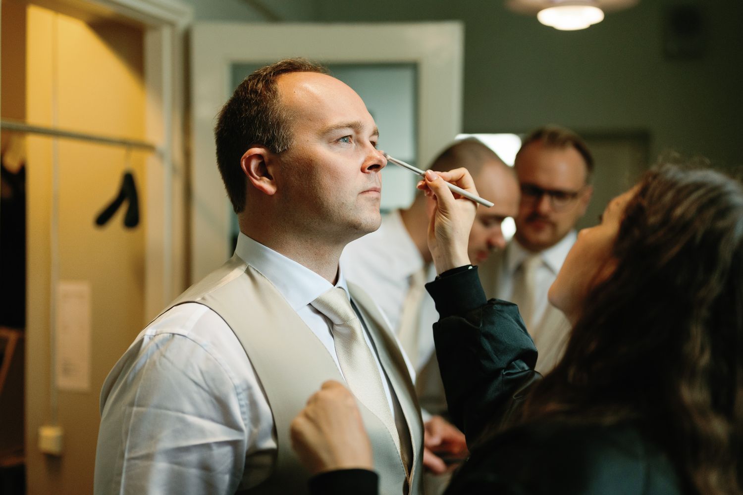 A makeup artist applies cosmetics to a man in a vest and tie, while another man stands in the background.