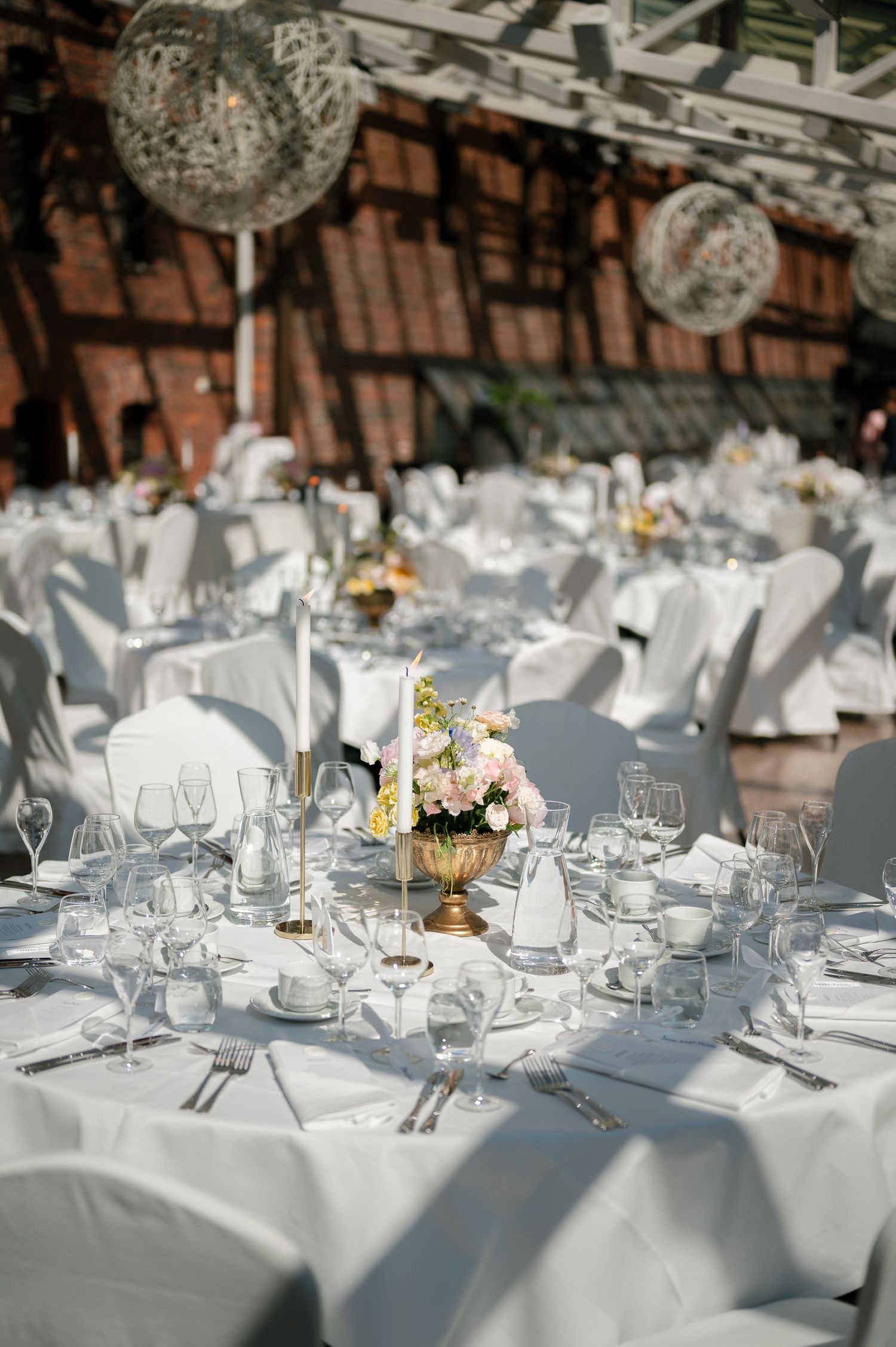 Elegant wedding reception tables with white linens, floral centerpieces, crystal glassware, and decorative wicker ball lights overhead.