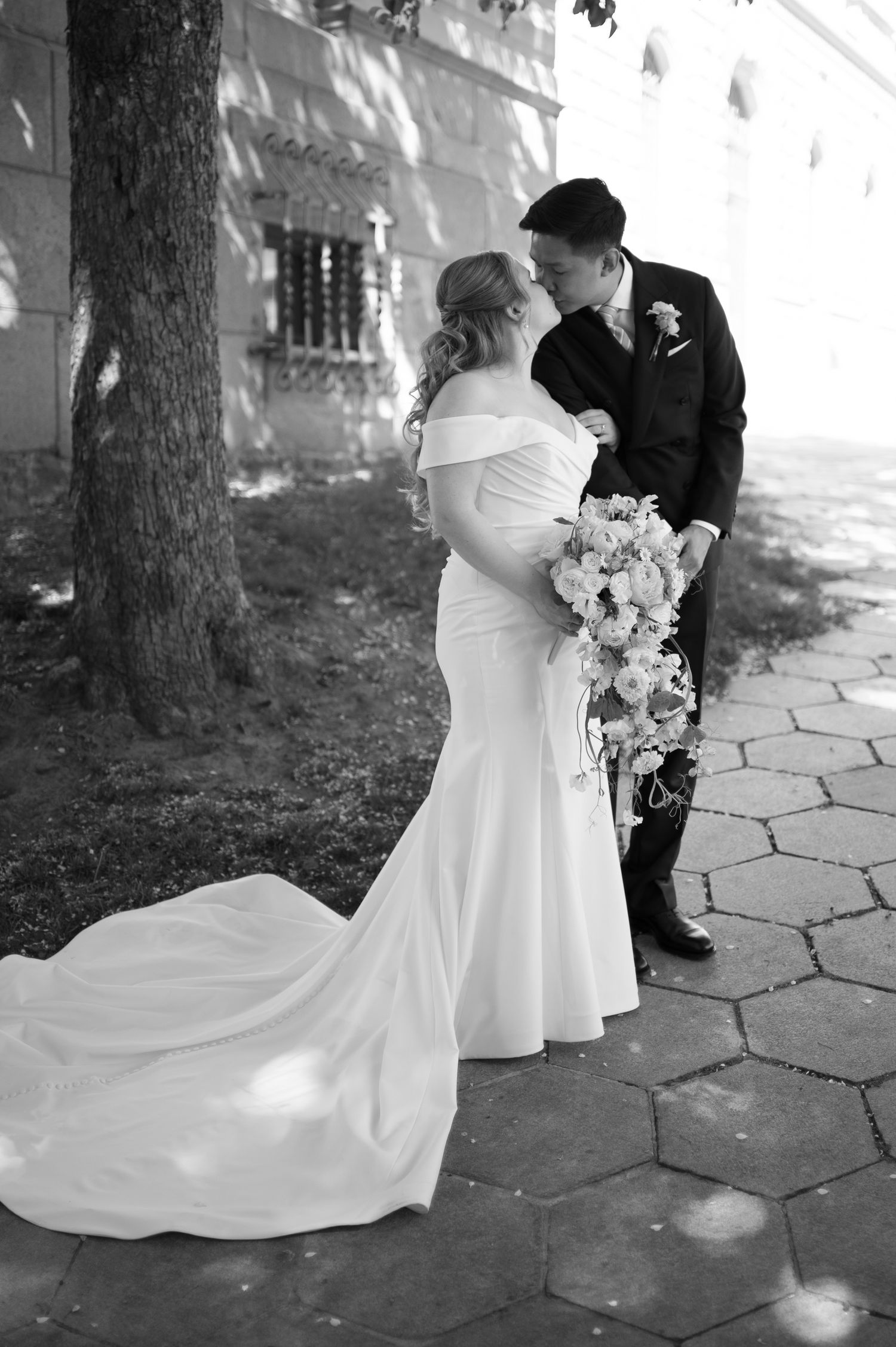 Black and white photo of bride and groom sharing a kiss outdoors, bride wearing elegant off-shoulder gown with long train.