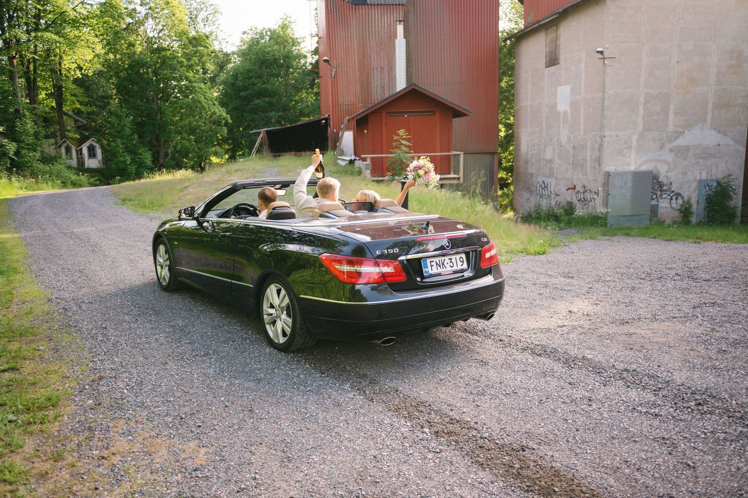 Black convertible car driving away on gravel path near rustic barn buildings.
