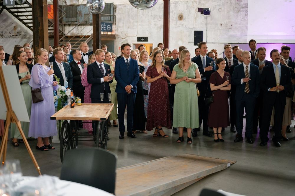 Wedding guests gathered for speeches and toasts in a converted warehouse venue with brick walls.