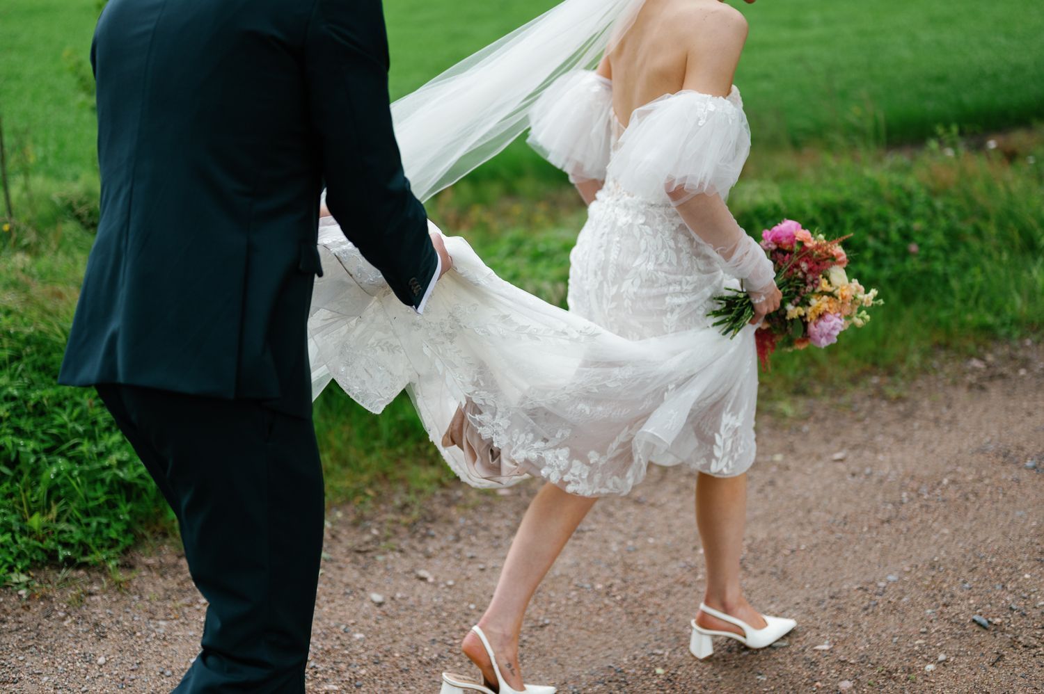 Detail shot of wedding couple's lower half walking on dirt path with bride holding bouquet.