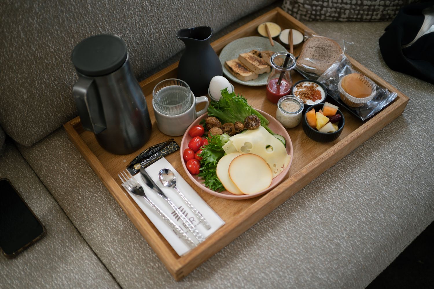 Wooden breakfast tray with fresh vegetables, cheese, toast, coffee, and a kettle served on a cozy sofa.