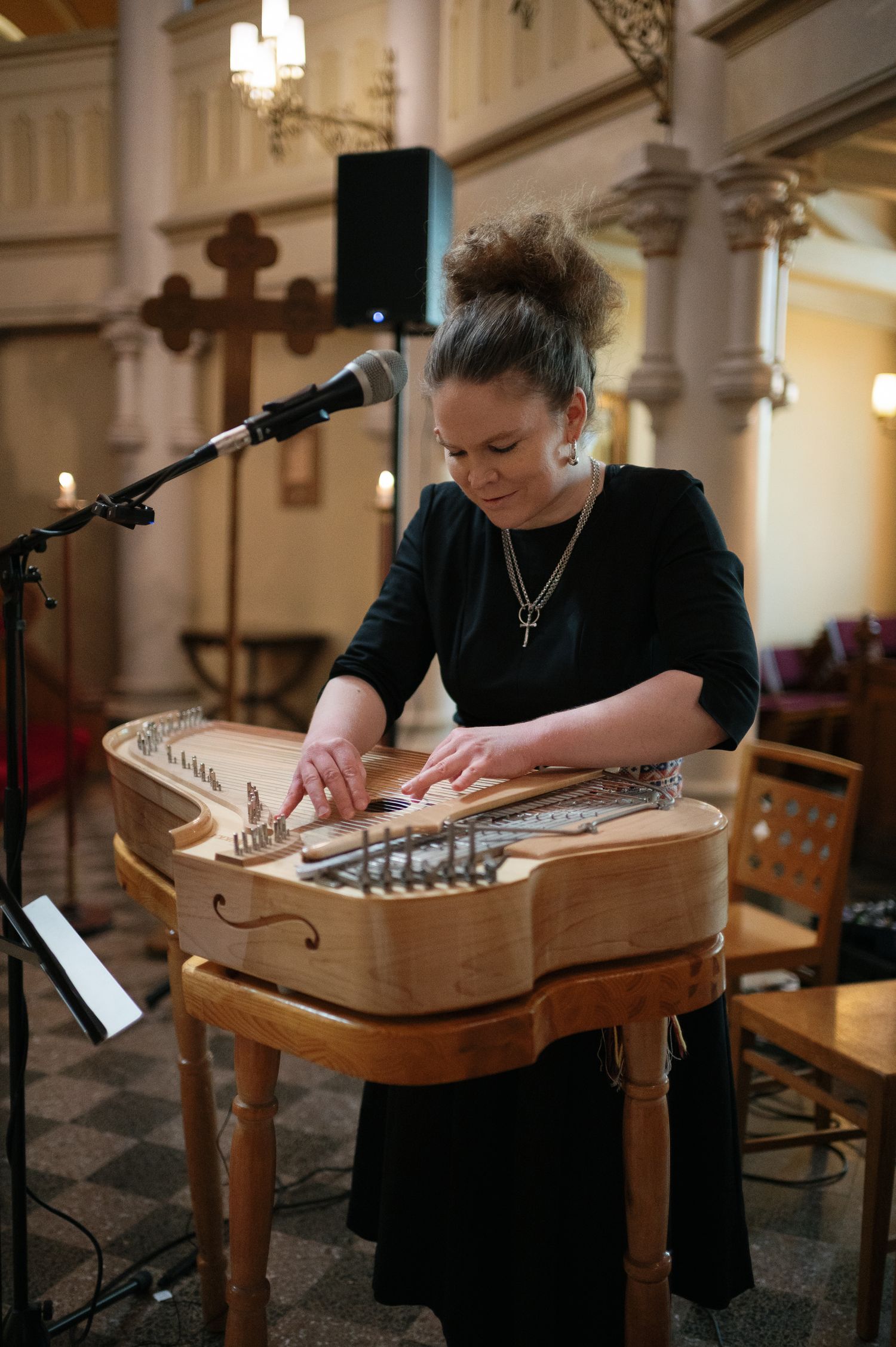 Young woman in black dress playing a nyckelharpa instrument in a church setting with a microphone stand nearby.