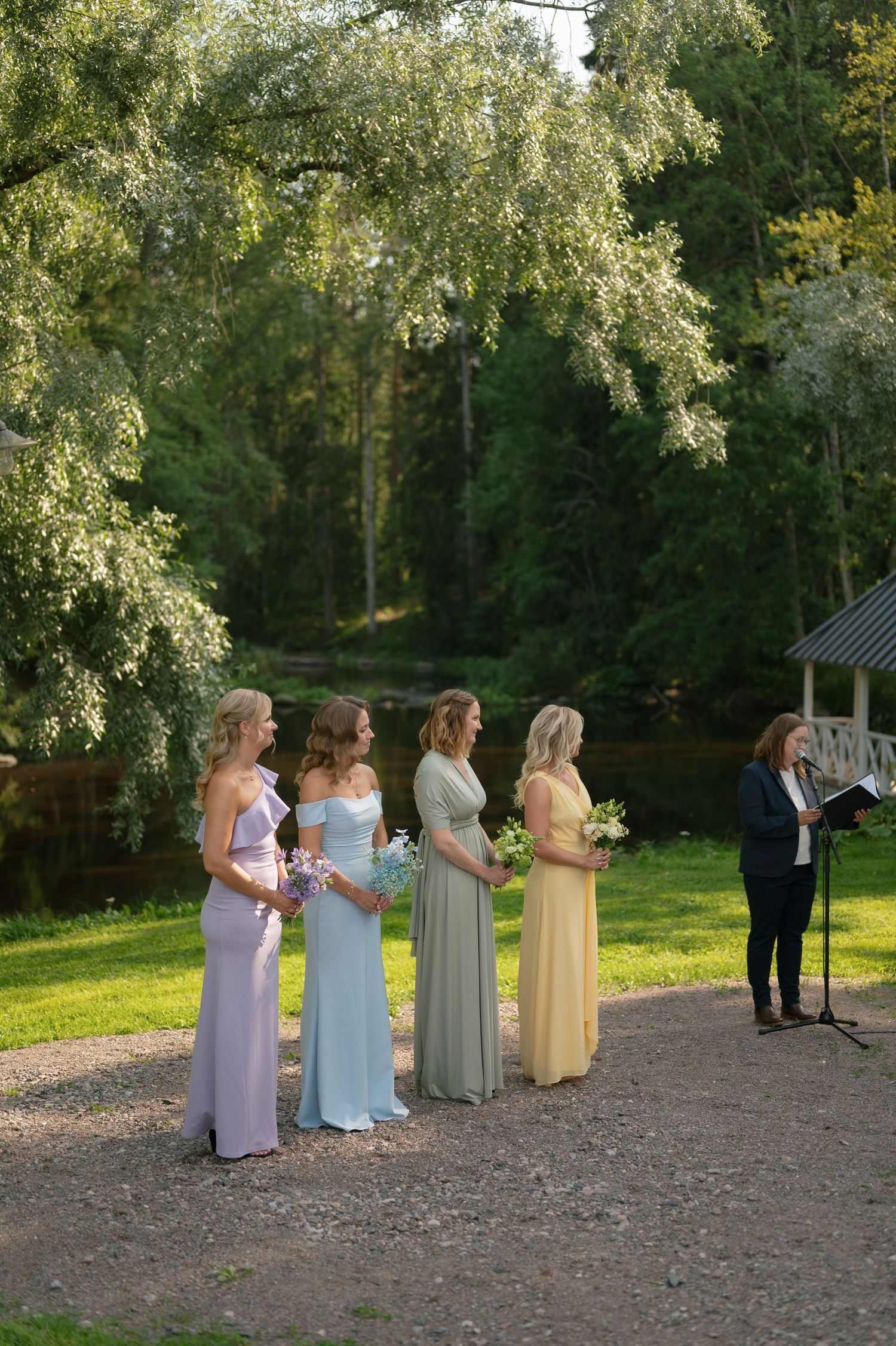 Bridesmaids in pastel dresses stand together during an outdoor wedding ceremony with trees in the background.