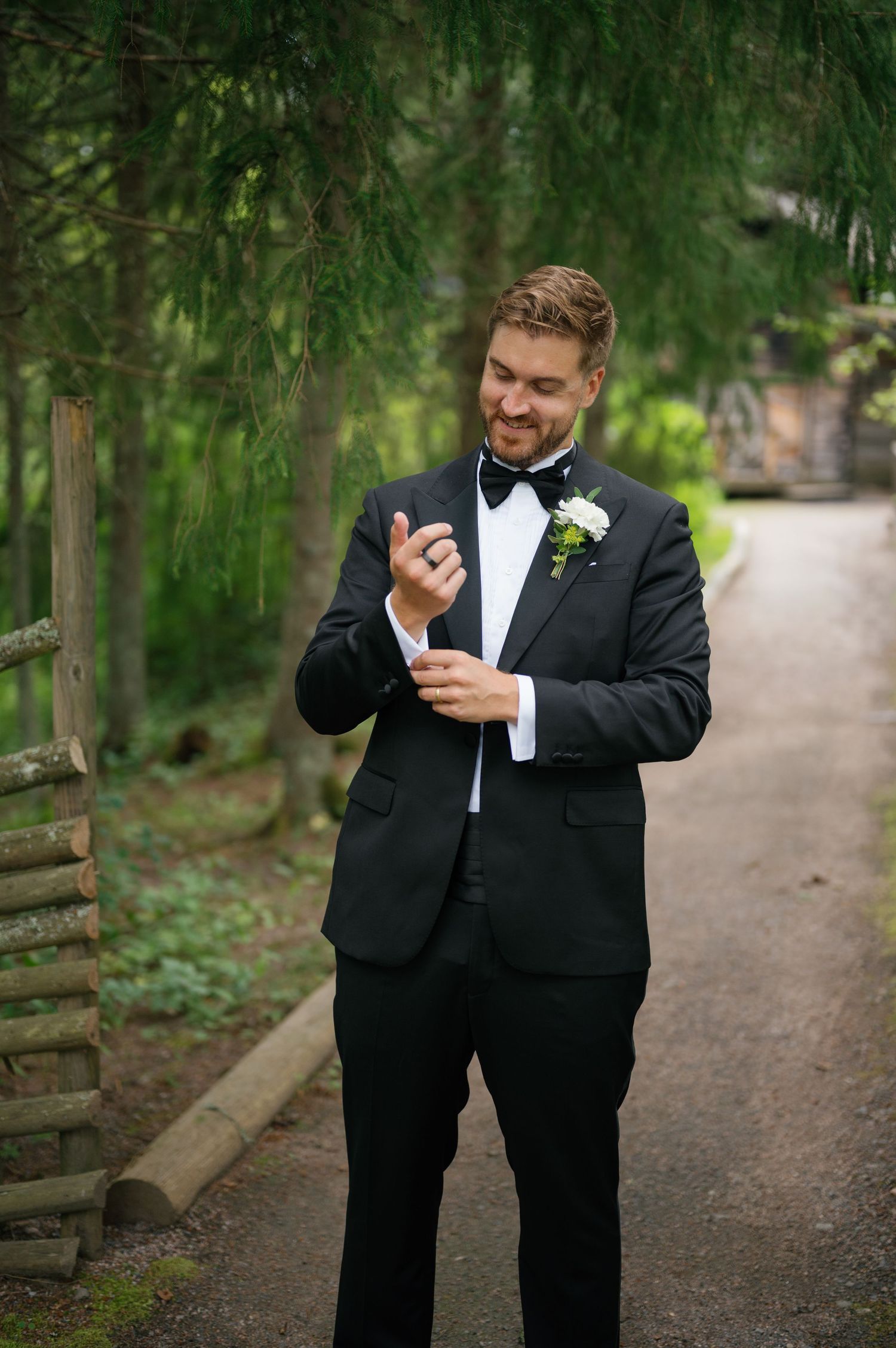 Two groomsmen in black tuxedos sharing a lighthearted moment on a tree-lined path.