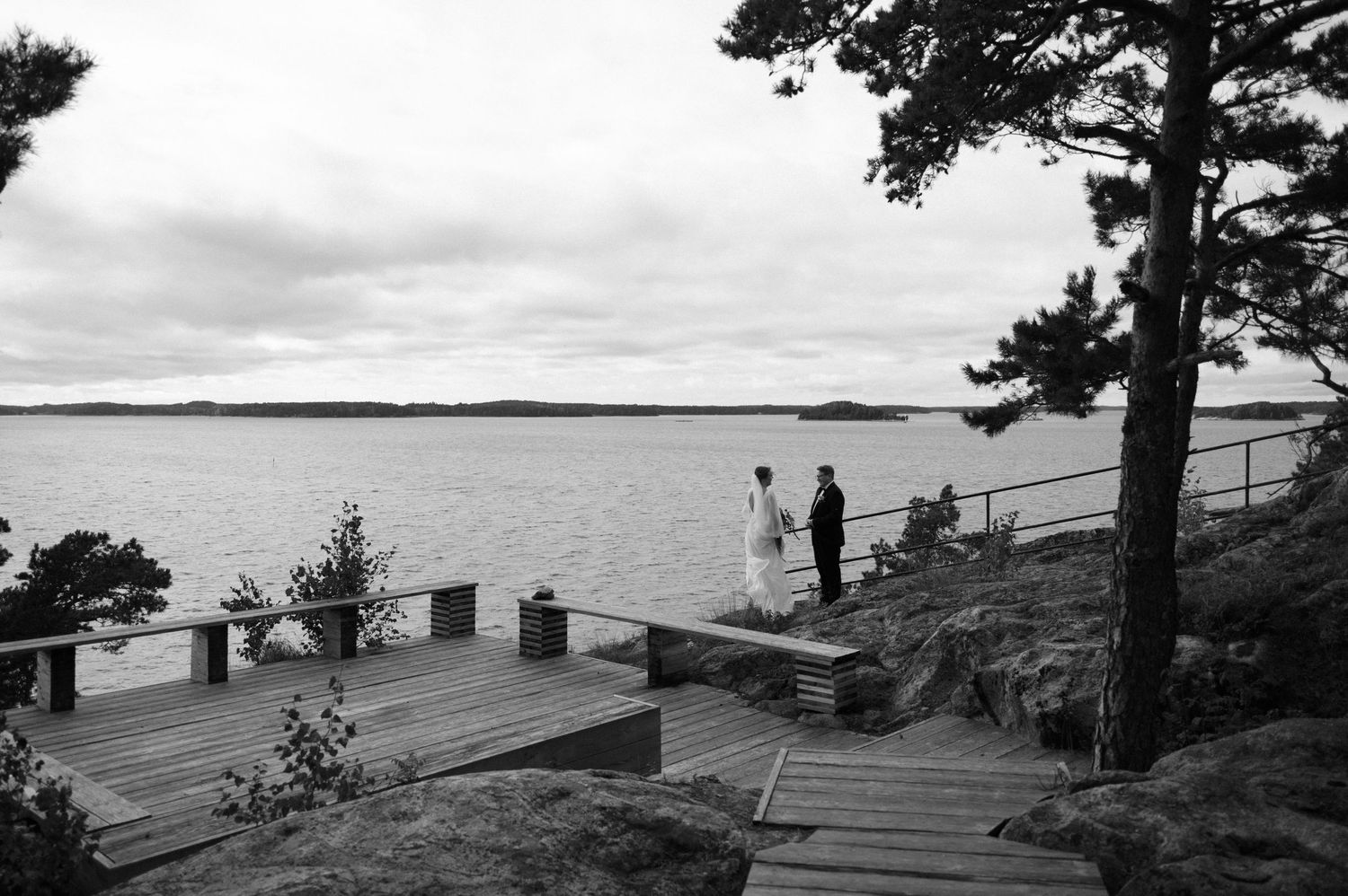 A black and white panoramic sequence of coastal landscapes with pine trees and wooden structures along a rocky shoreline.
