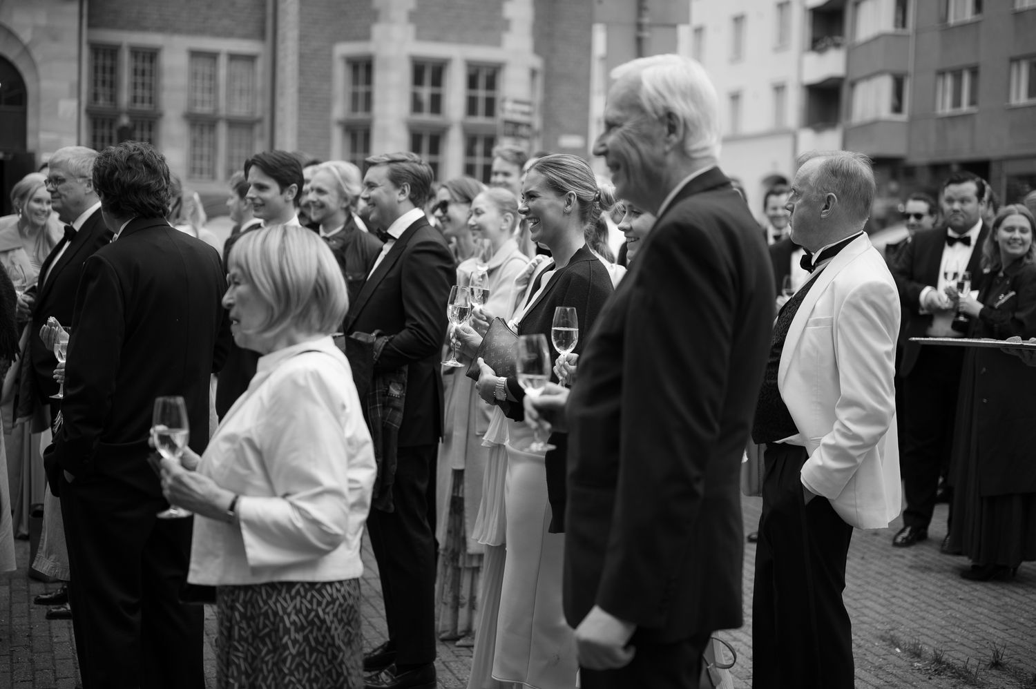 A black and white photograph shows a crowd of people gathered outside on a city street during a social event.
