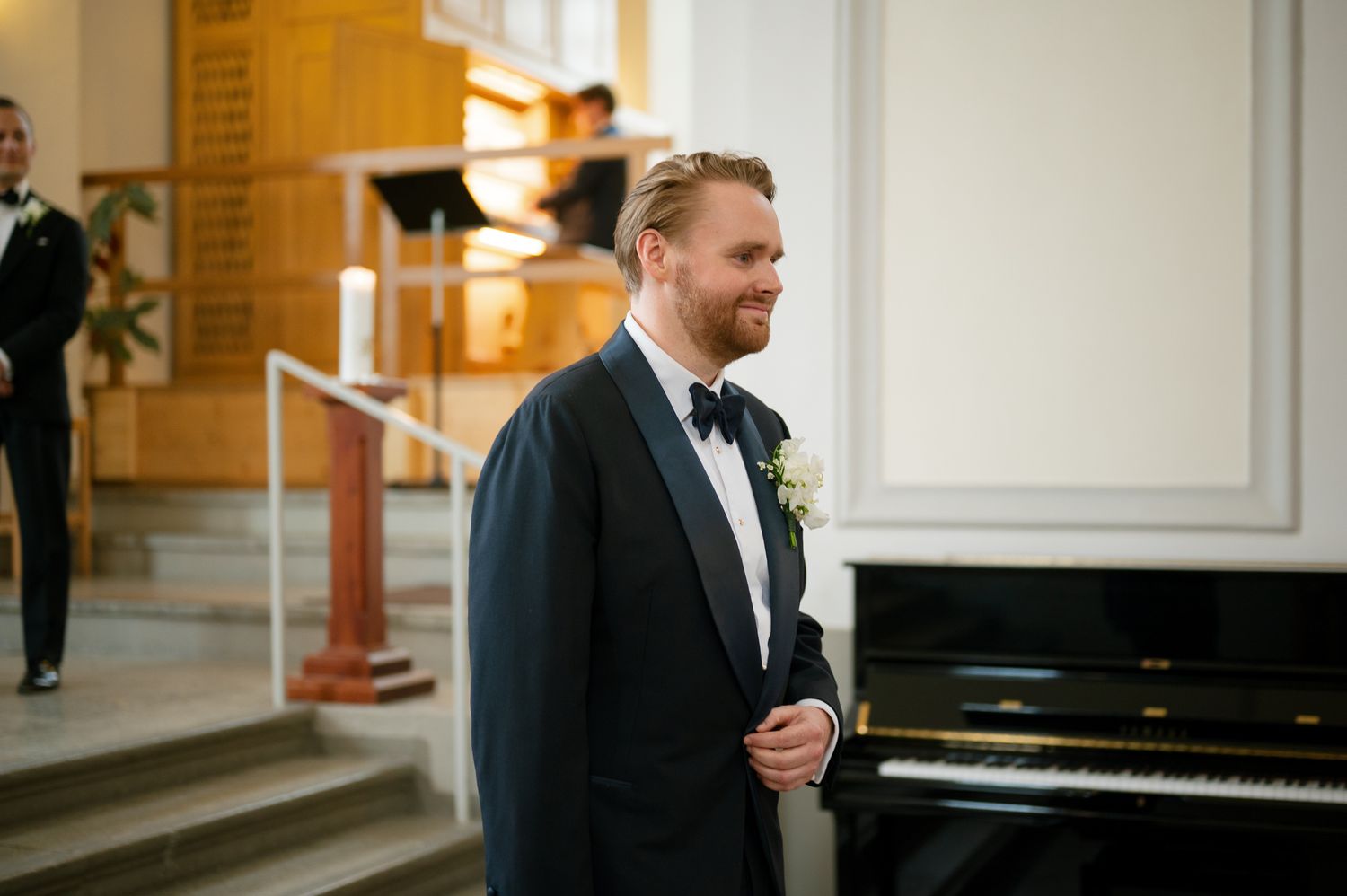 A person in a black tuxedo stands next to a piano in an elegant room with white walls and architectural details.
