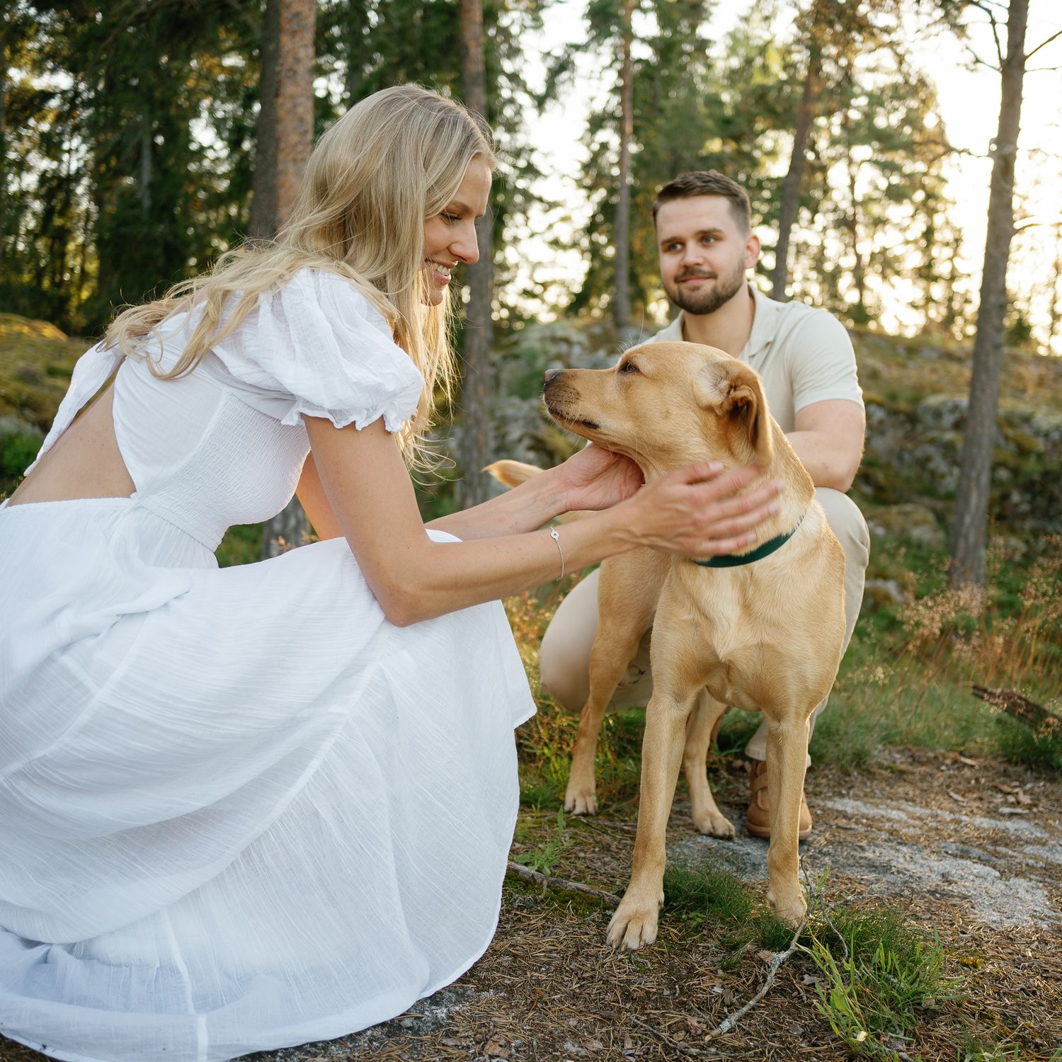 A couple in white clothing pets a friendly yellow dog in a forest setting.