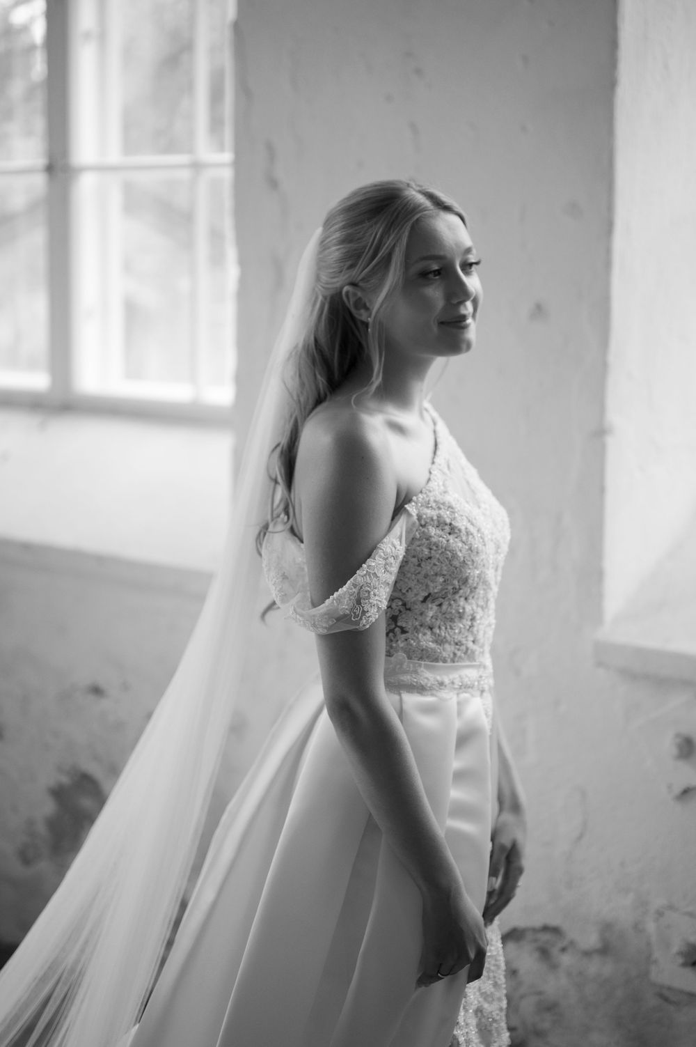 Black and white portrait of a bride in a flowing veil standing by bright windows.
