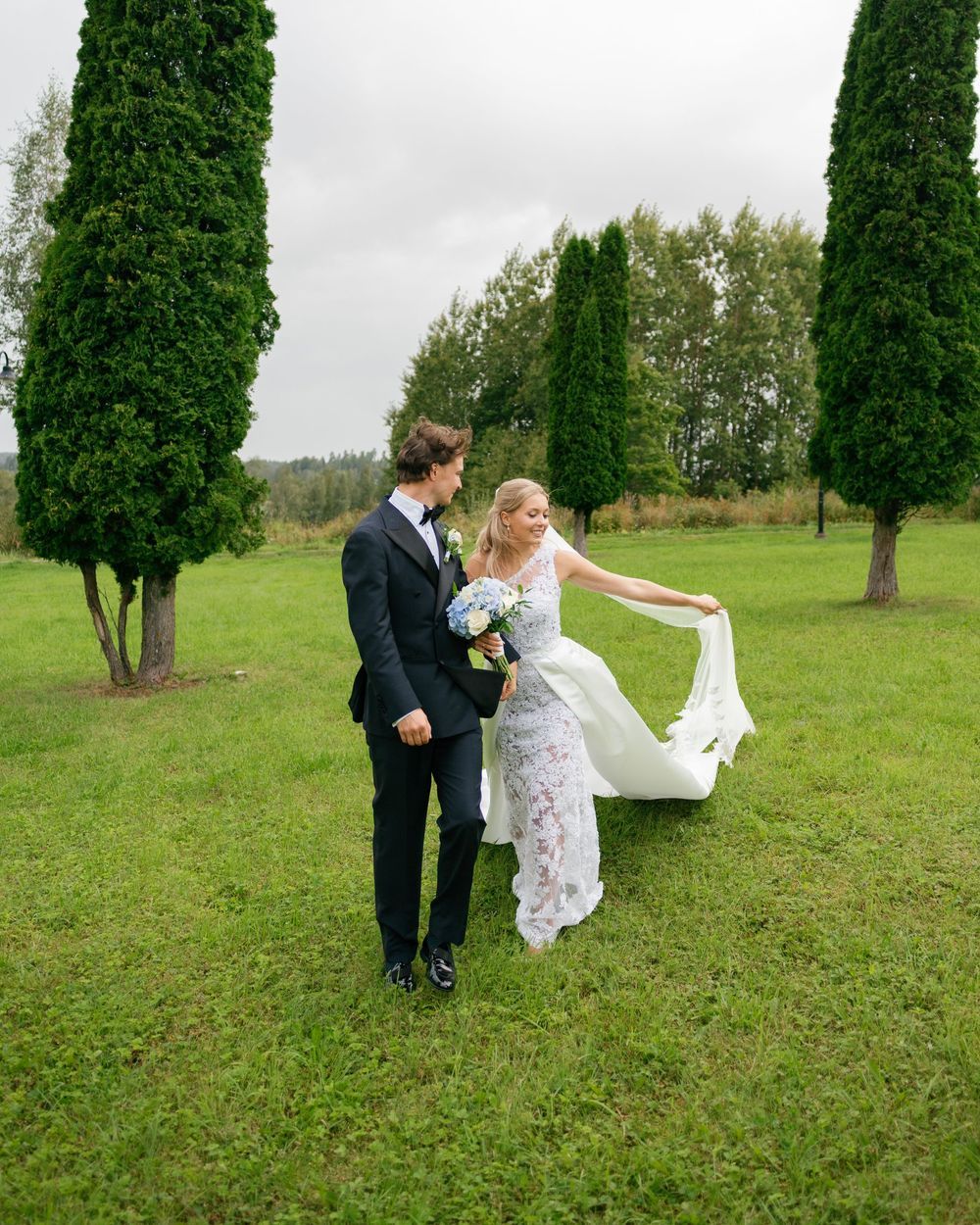 Newlyweds playfully interact on a grassy hill with cypress trees.