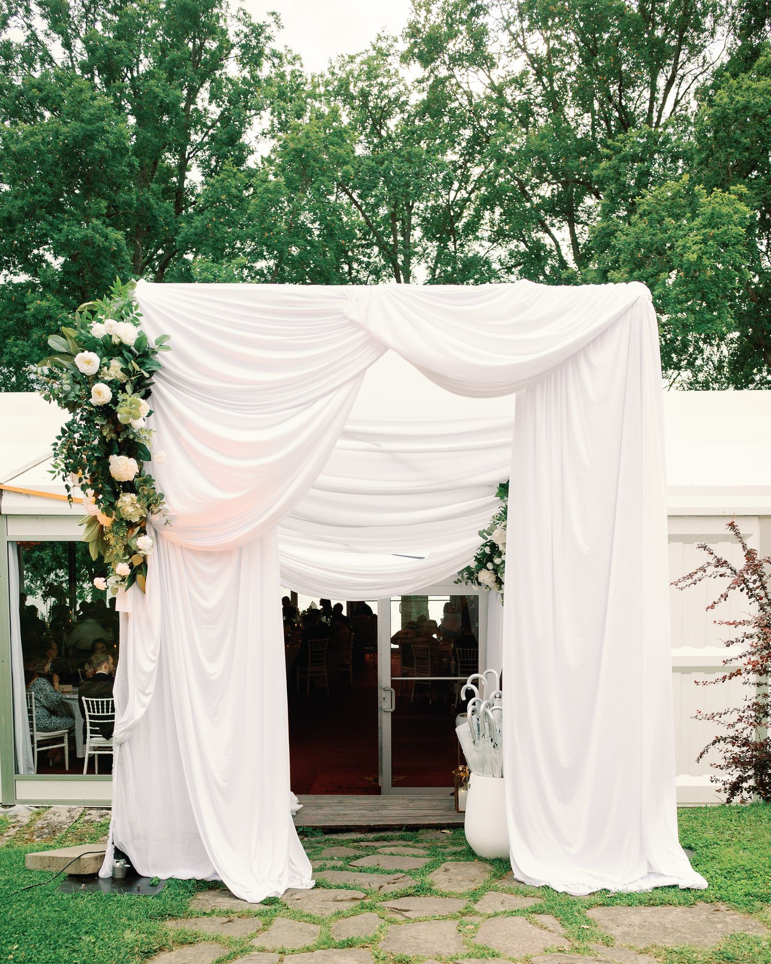 Elegant outdoor wedding arch draped in white fabric with floral arrangements, set against lush green trees.