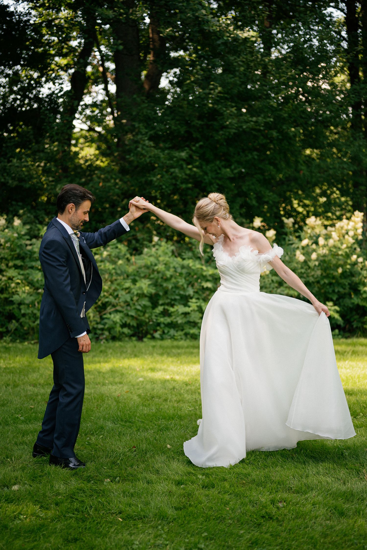 Bride in white gown twirls as groom holds her hand in a lush green garden during outdoor wedding celebration.