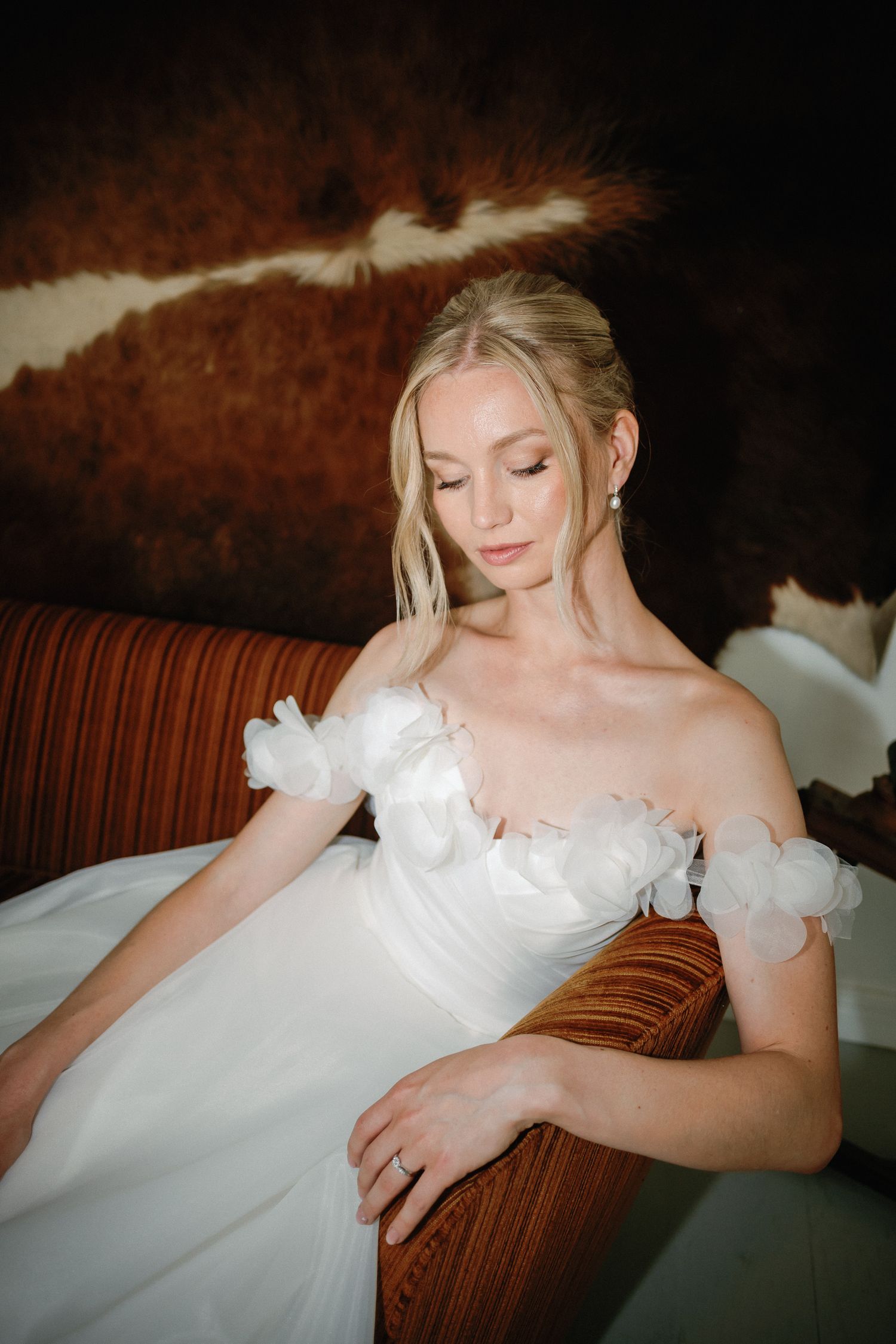 Elegant bride in white off-shoulder gown with 3D floral accents sits gracefully on a brown velvet sofa.