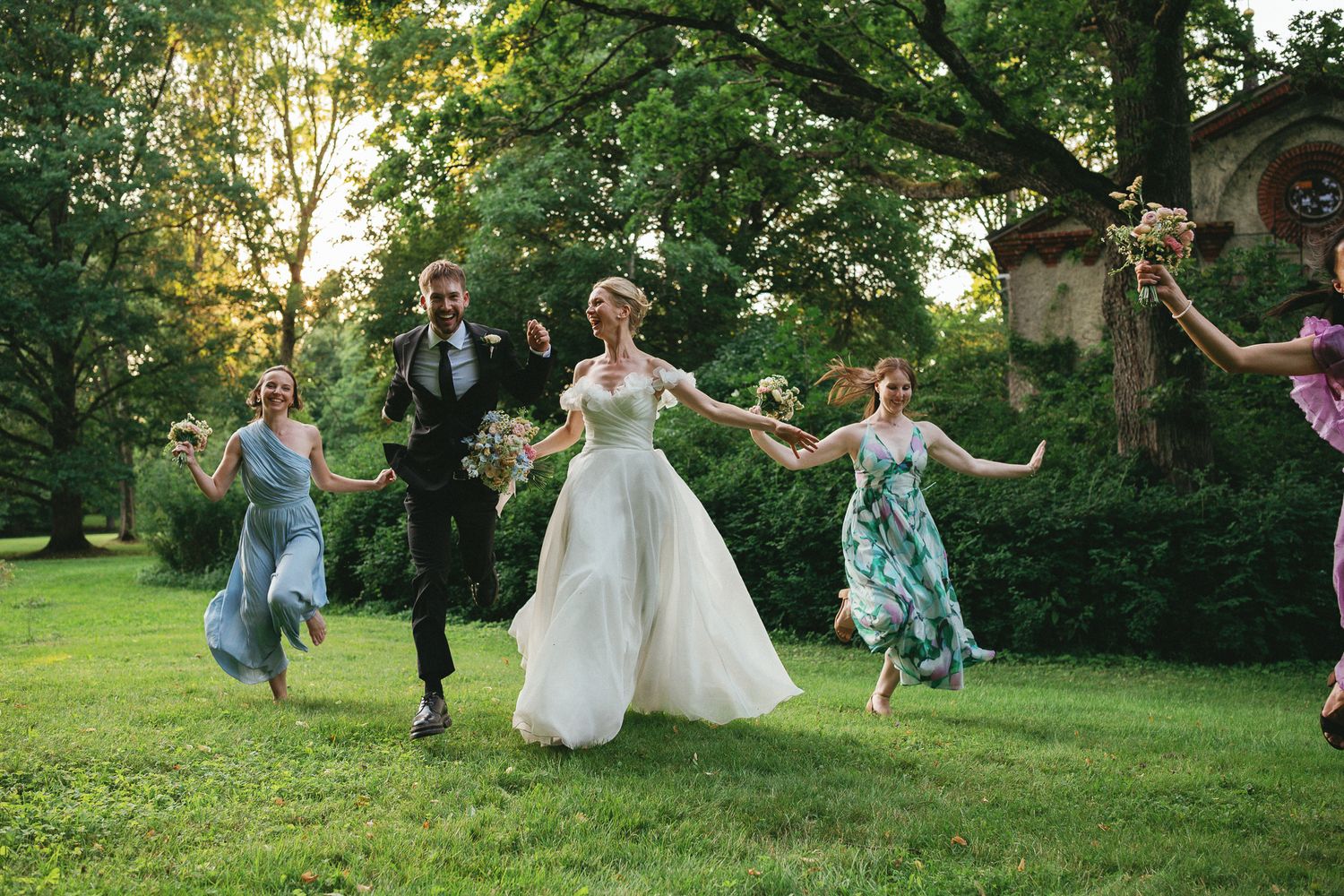 Joyful wedding party running on green lawn with bride in white gown, groom, and bridesmaids in colorful dresses outdoors.