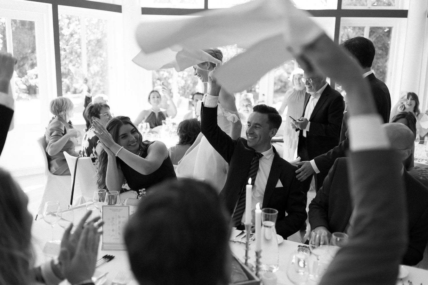 Black and white wedding reception photo of guests joyfully waving napkins at a decorated dining table.