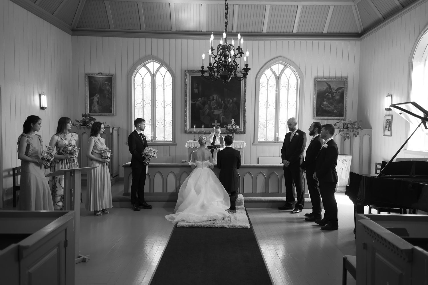 Black and white wedding ceremony in a chapel with bridesmaids, groomsmen, and couple at the altar.