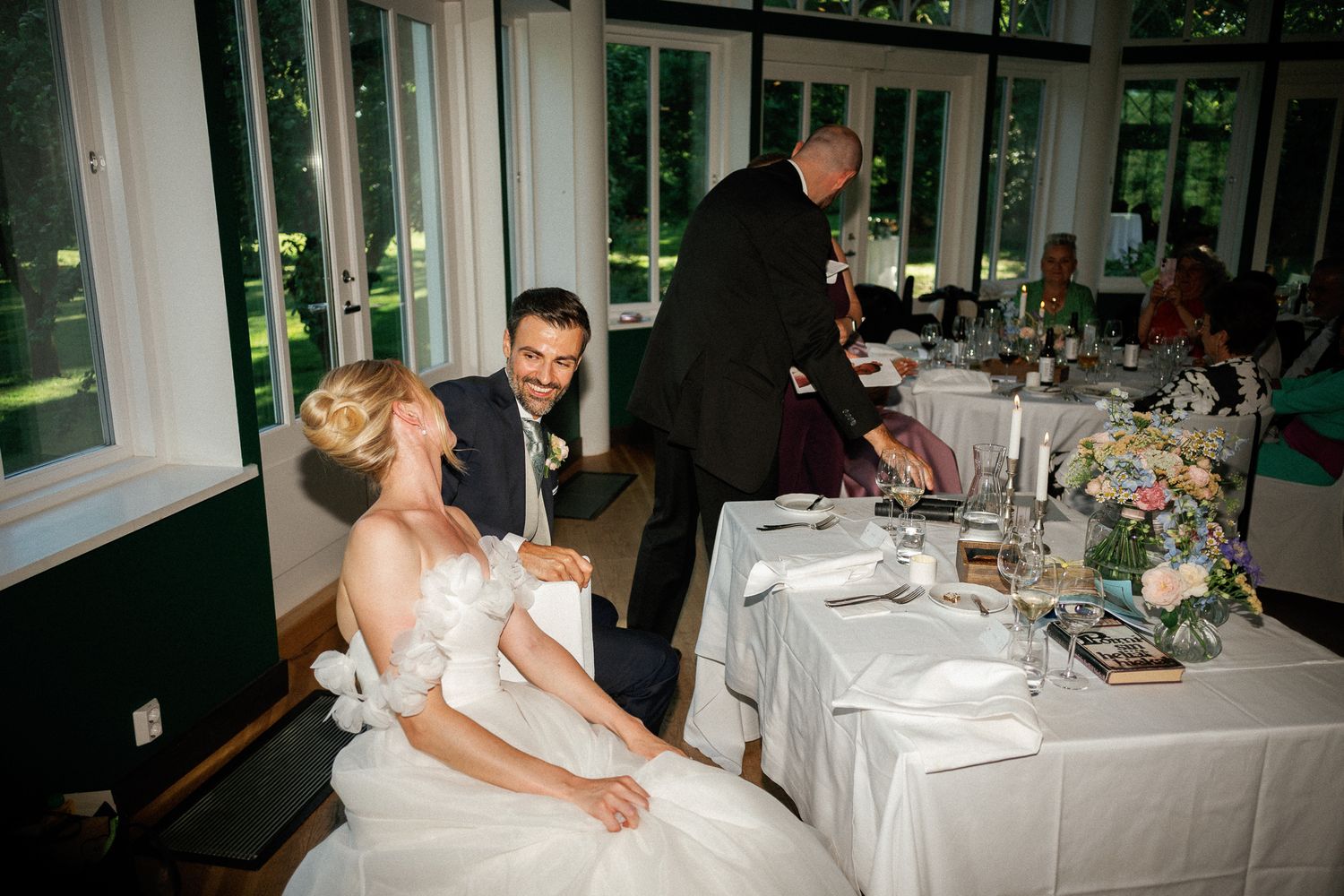 Bride and groom share a joyful moment at their elegant wedding reception table, with guests and floral centerpieces nearby.