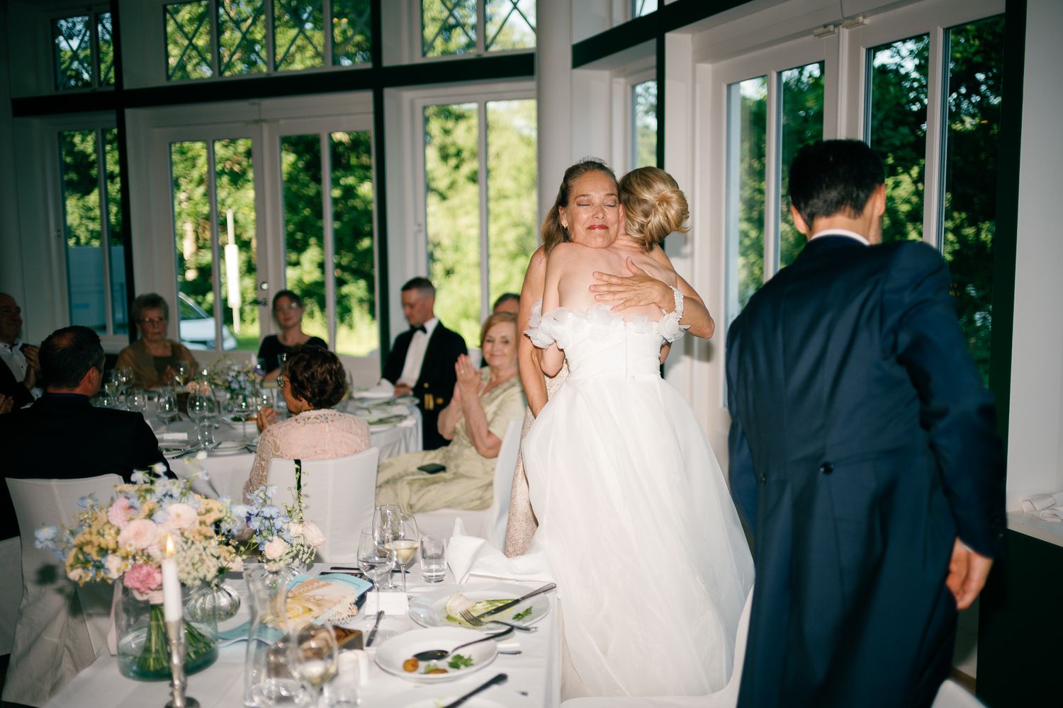 Bride in white gown hugging a guest at an elegant wedding reception with floral centerpieces and large windows.