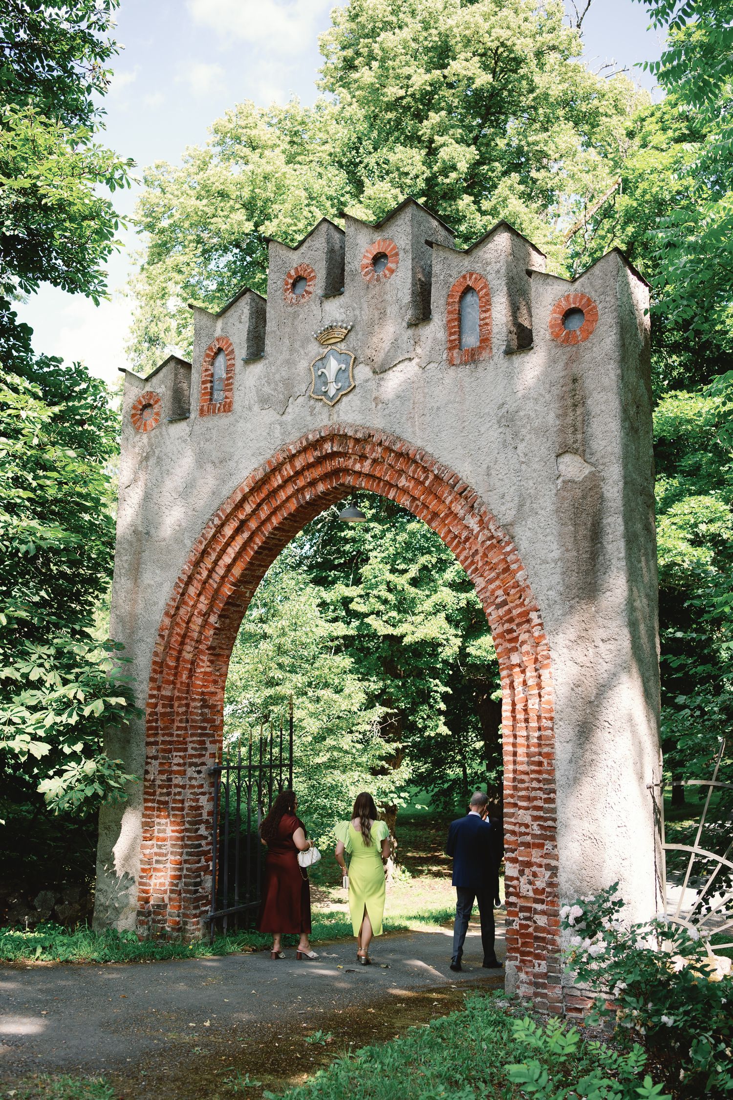 People walking through a historic red brick Gothic arch gateway surrounded by lush green trees in summer.