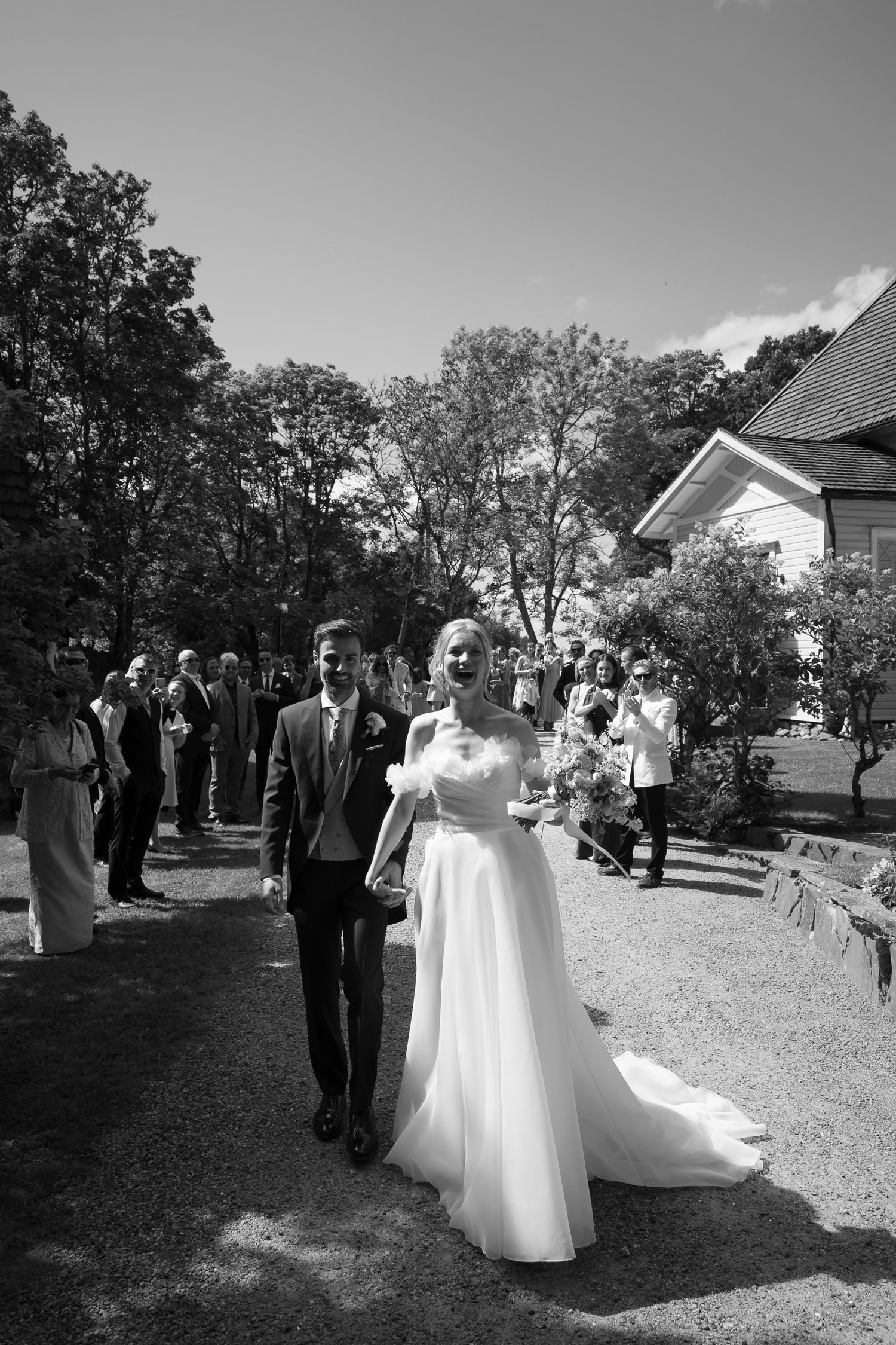 Black and white photo of a smiling bride in a white gown and groom walking down an outdoor aisle with guests.