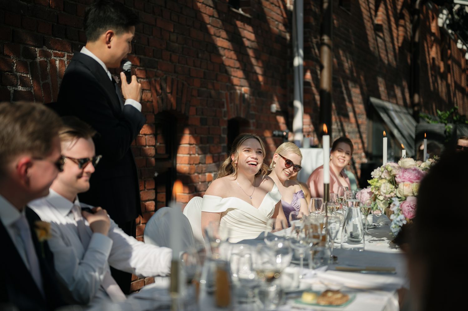 Bride laughing joyfully at outdoor wedding reception while groom gives a heartfelt speech to seated guests.