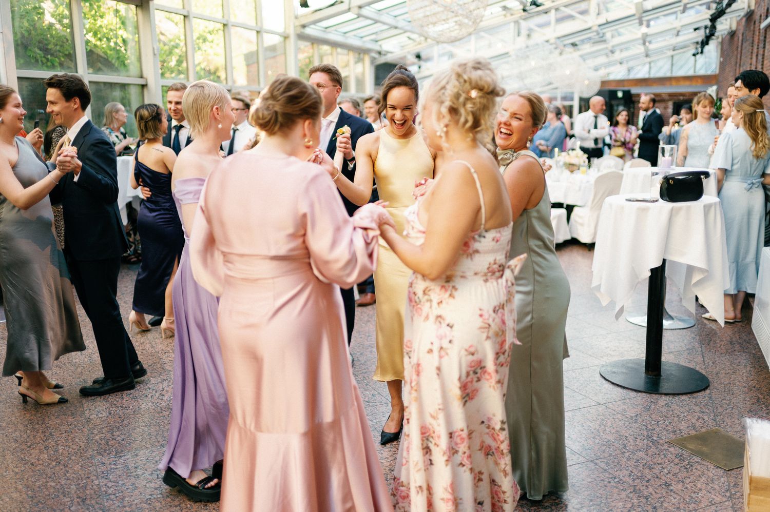 Guests in elegant gowns dancing together at a wedding reception in a bright, modern venue.