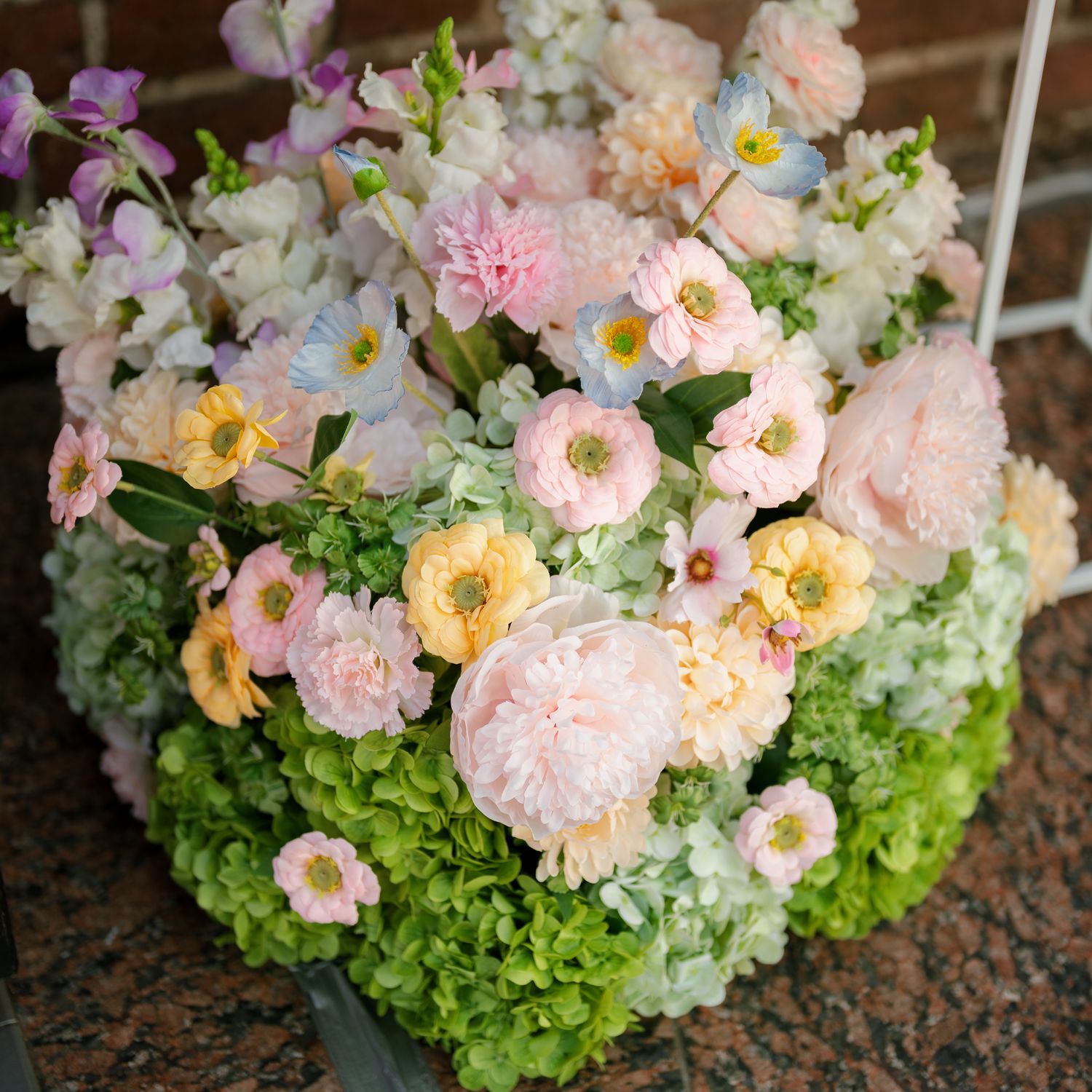 Lush floral arrangement featuring pink peonies, yellow ranunculus, blue anemones, and green hydrangeas in a pastel spring bouquet.