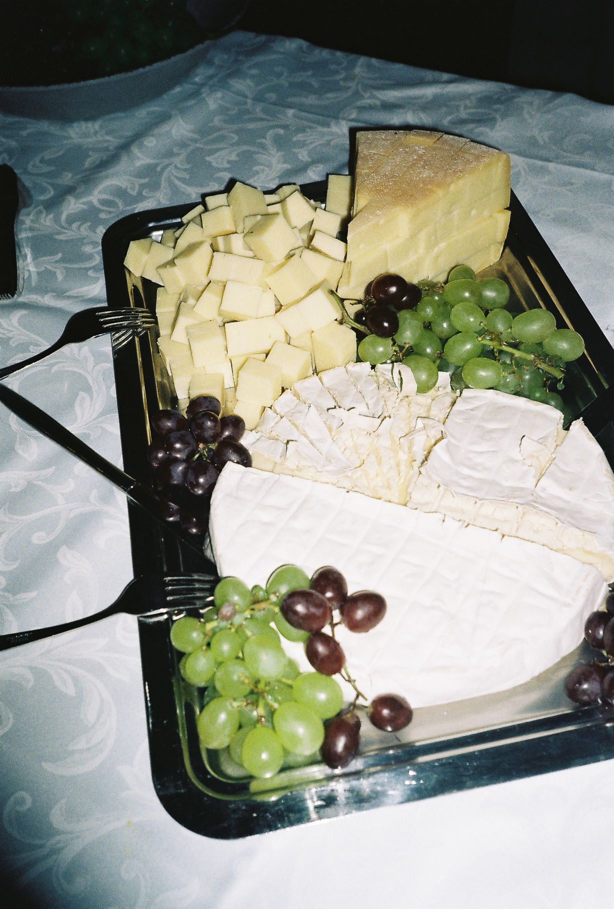 A black tray with assorted cheeses including brie and cubed varieties, garnished with green and red grapes on a white tablecloth.
