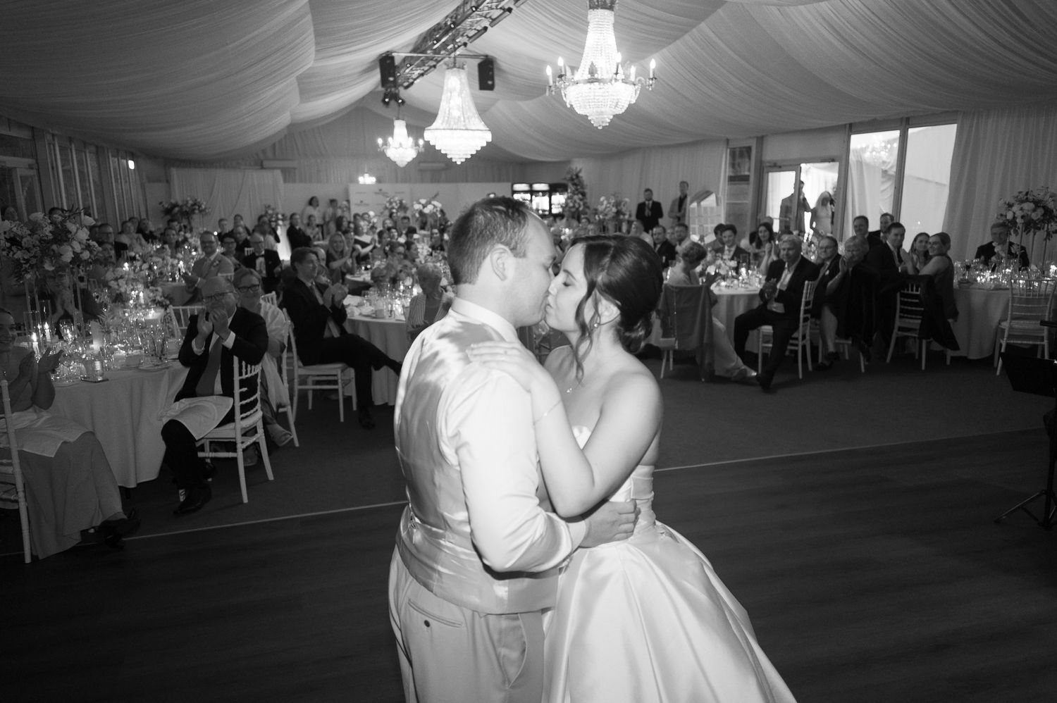 Black and white photo of a couple sharing their first dance kiss at a wedding reception with guests seated at tables.