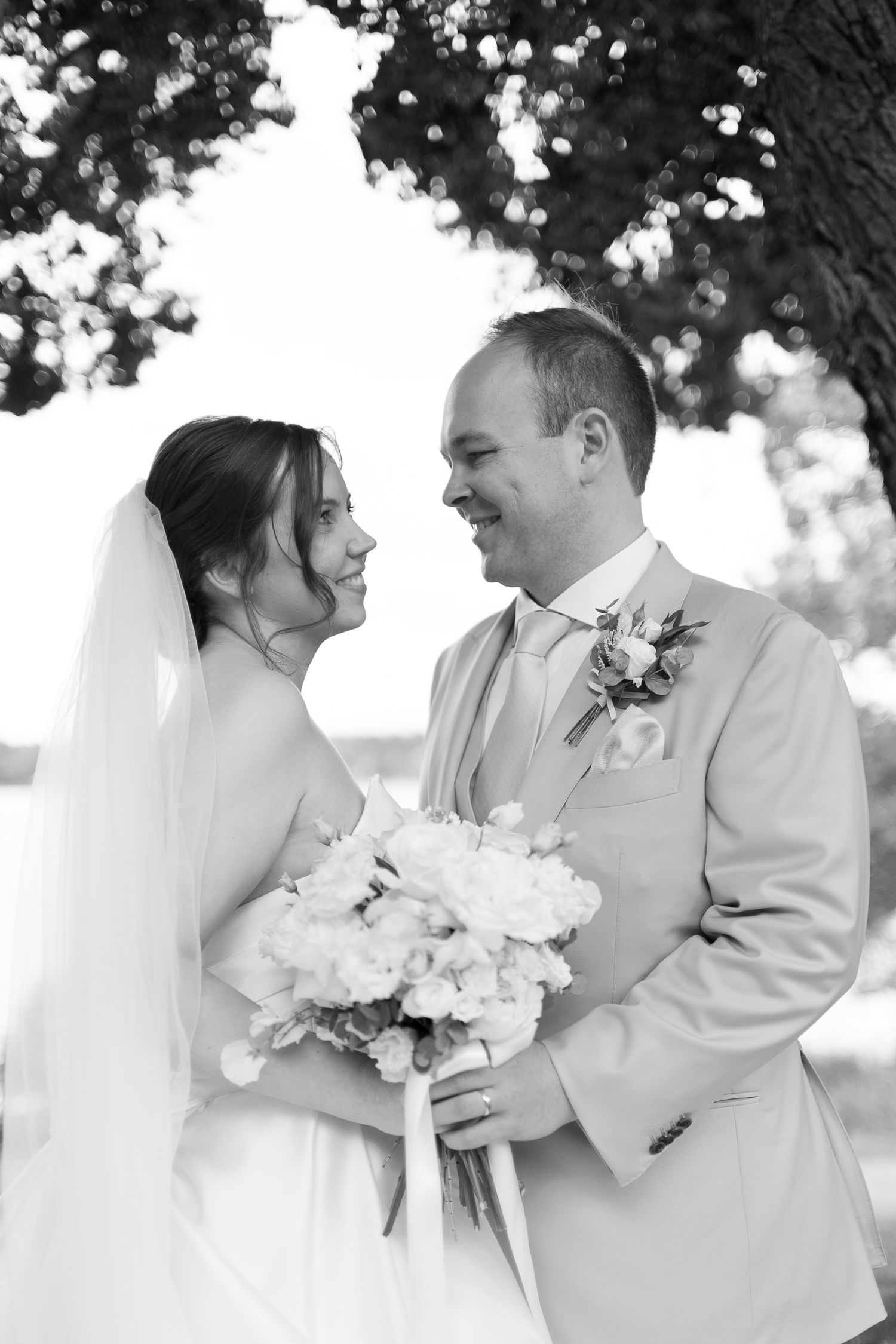 Black and white photo of smiling bride and groom holding bouquet, standing under a tree on their wedding day.