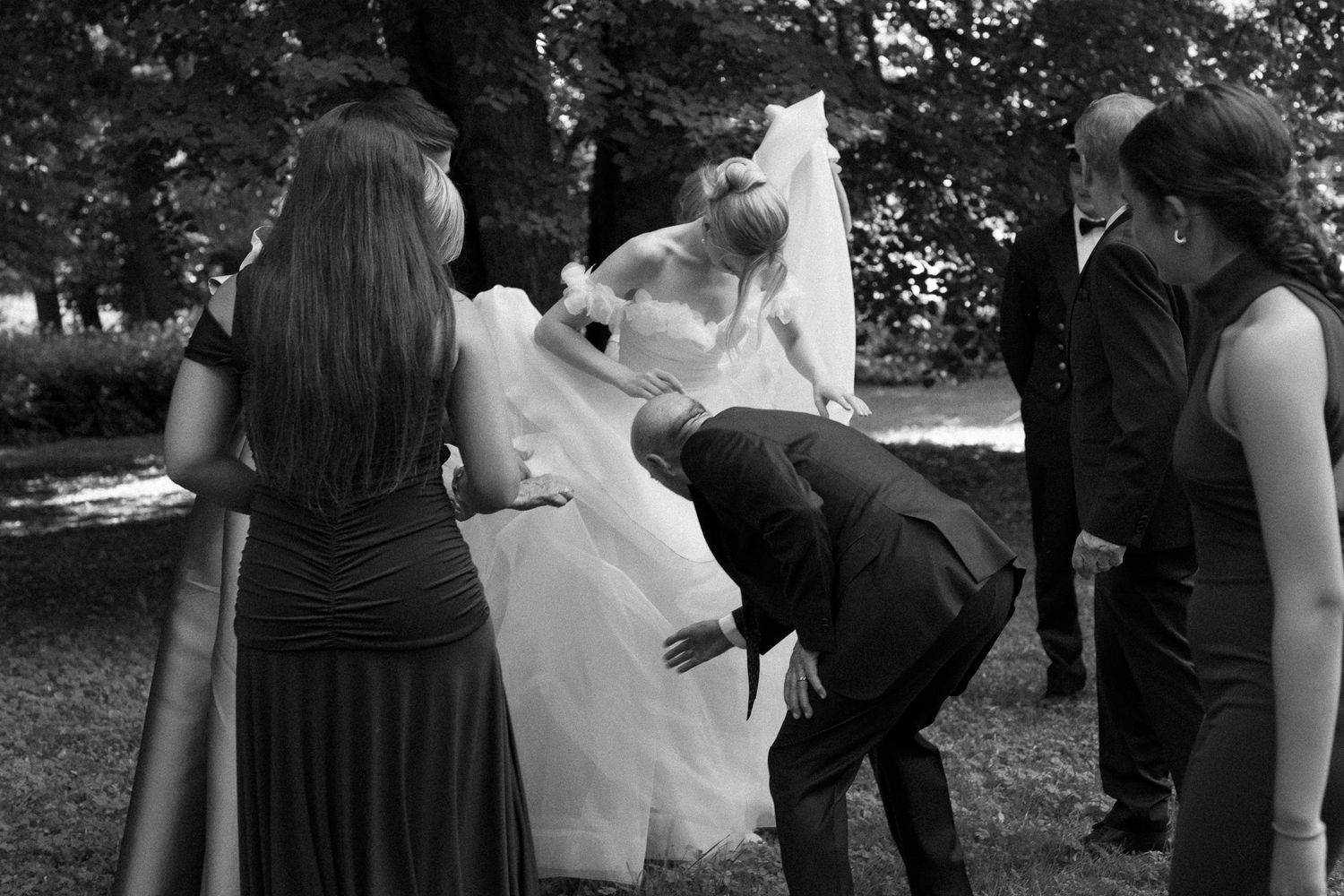 Black and white wedding photo of bride in white gown and veil surrounded by guests outdoors near trees.