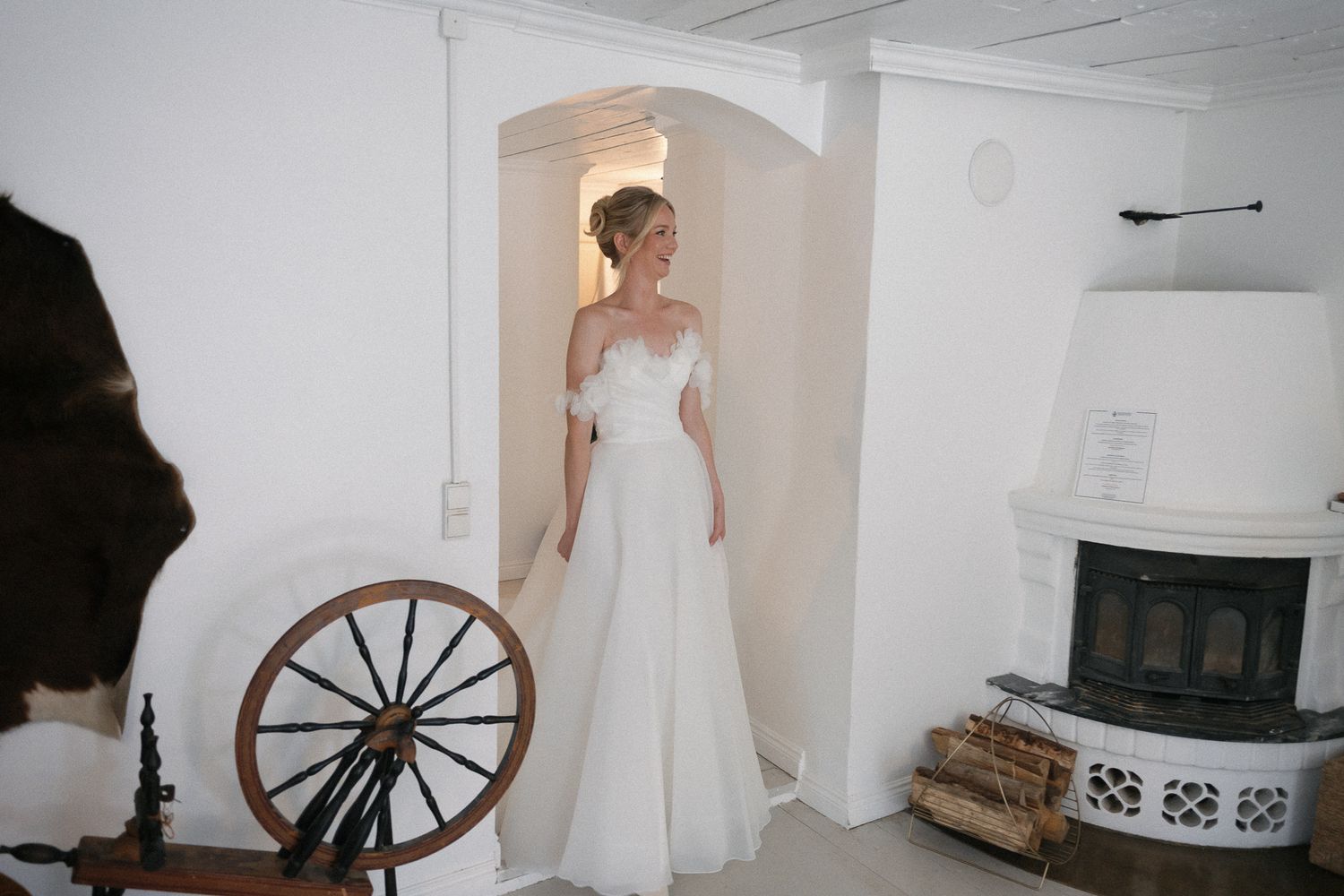 Bride in off-shoulder white wedding gown standing in archway beside vintage spinning wheel and fireplace.