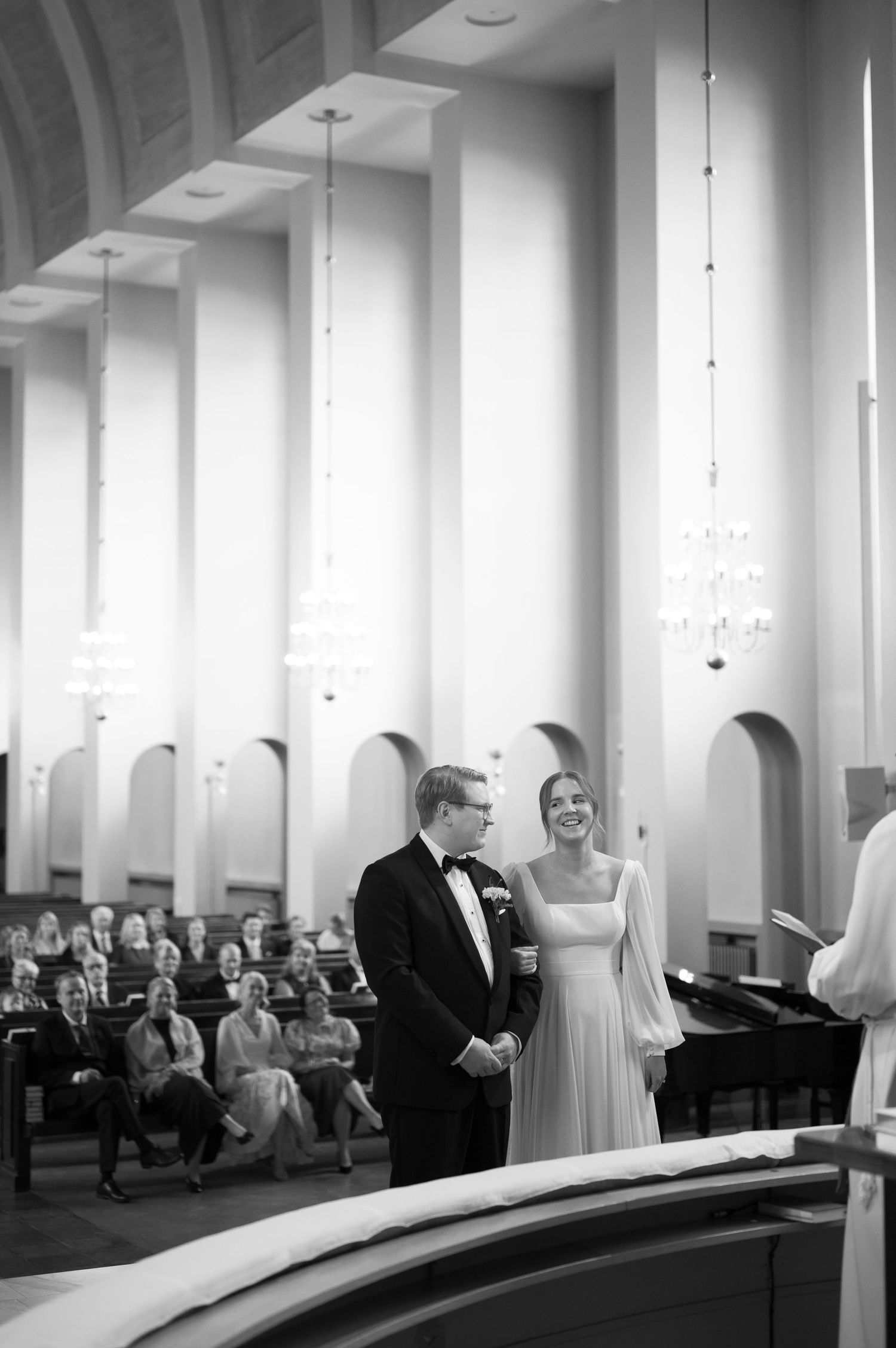 Black and white panoramic photo of a wedding ceremony taking place in a modern church with white columns and pews.