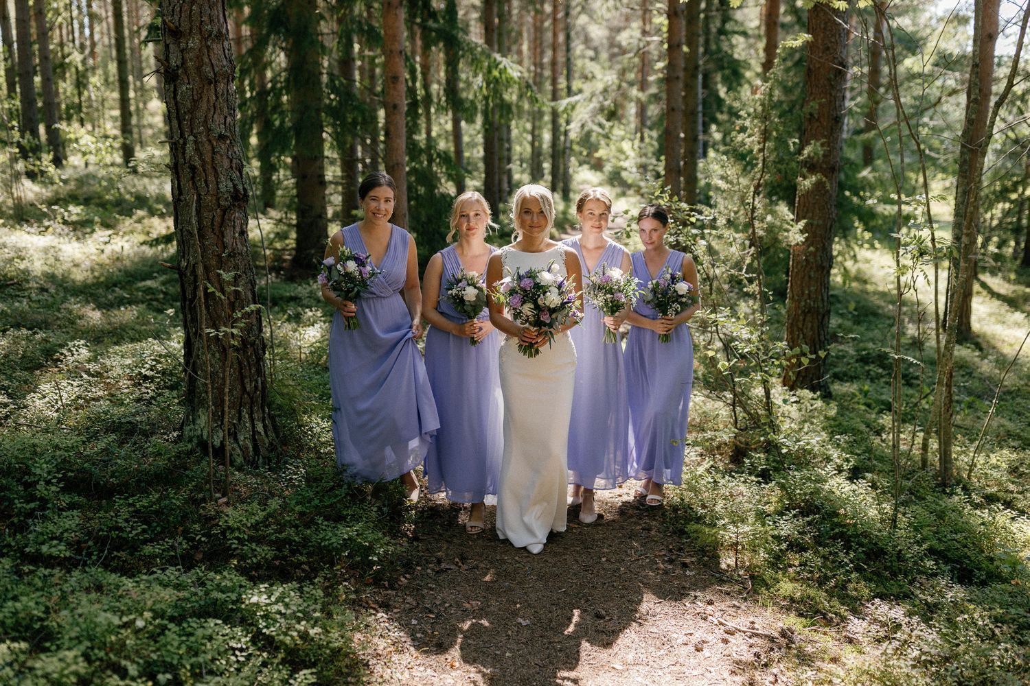Wedding party poses together among tall trees in a sun-dappled forest.