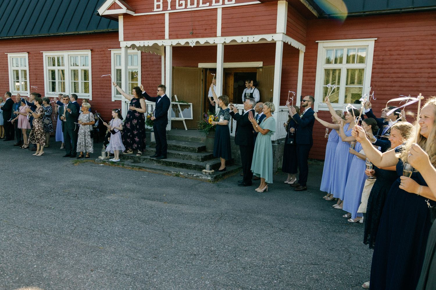 Wedding guests gather outside a red wooden building with green roof.