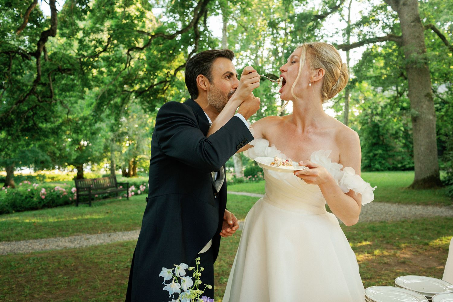 Bride and groom feeding each other wedding cake outdoors in a lush green garden setting.