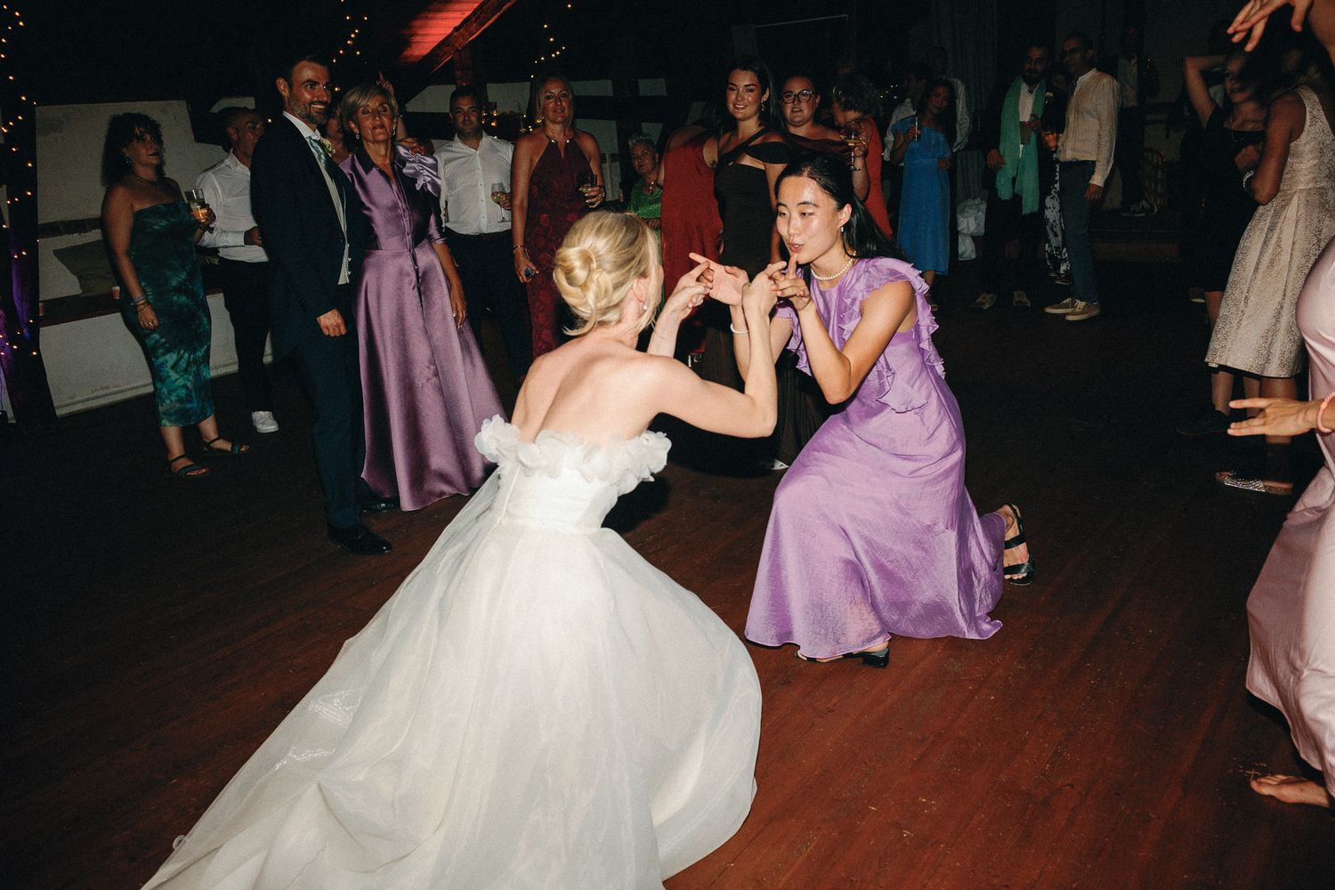 Bride in white gown dancing with bridesmaid in lavender dress on wedding reception dance floor.