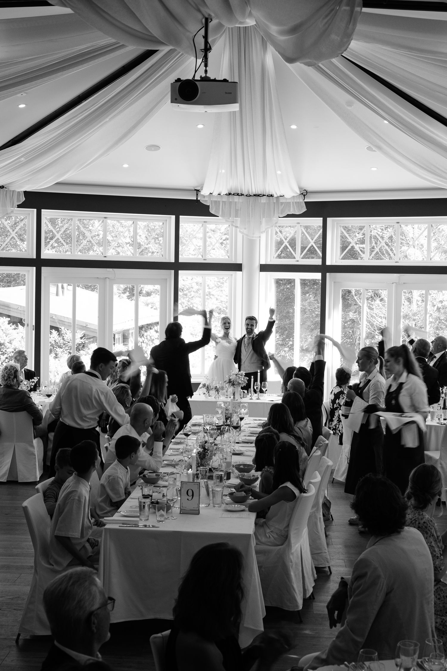 Black and white wedding reception photo showing bride and groom toasting guests in elegant draped venue.