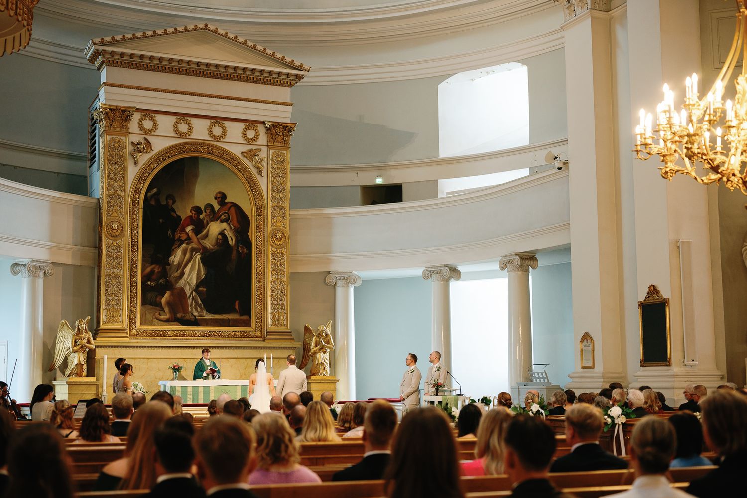 Elegant church ceremony with ornate golden altar, large religious painting, and congregation seated in wooden pews.