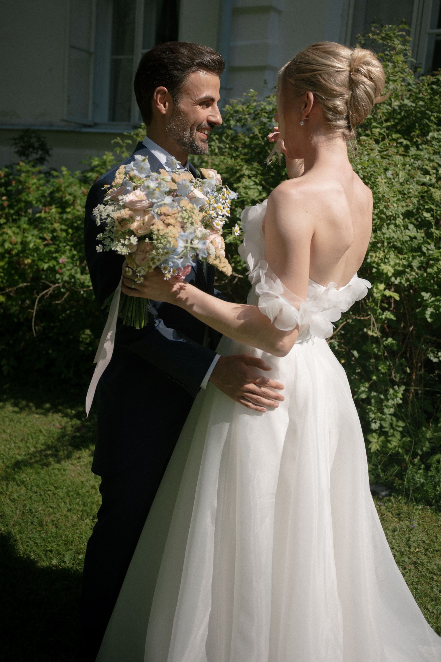 Smiling groom in black suit holds floral bouquet while embracing bride in elegant backless white tulle wedding gown outdoors.