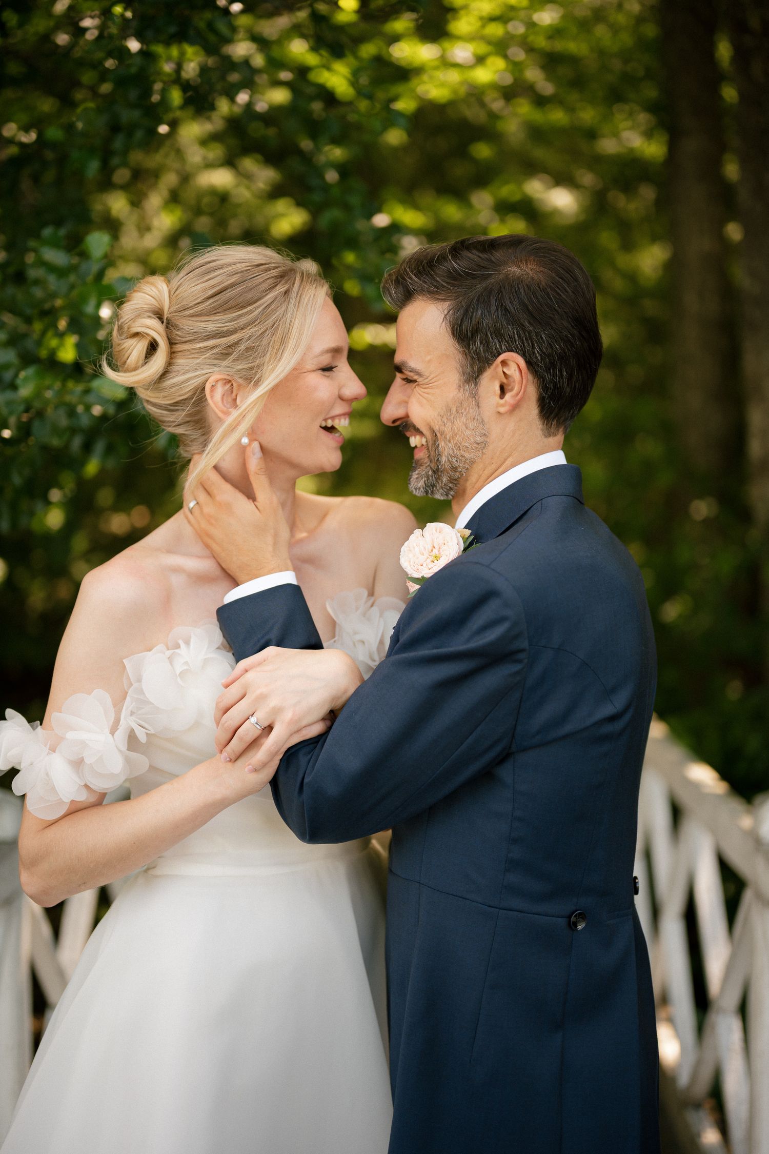 A smiling bride and groom in wedding attire share a romantic moment outdoors surrounded by lush green trees.