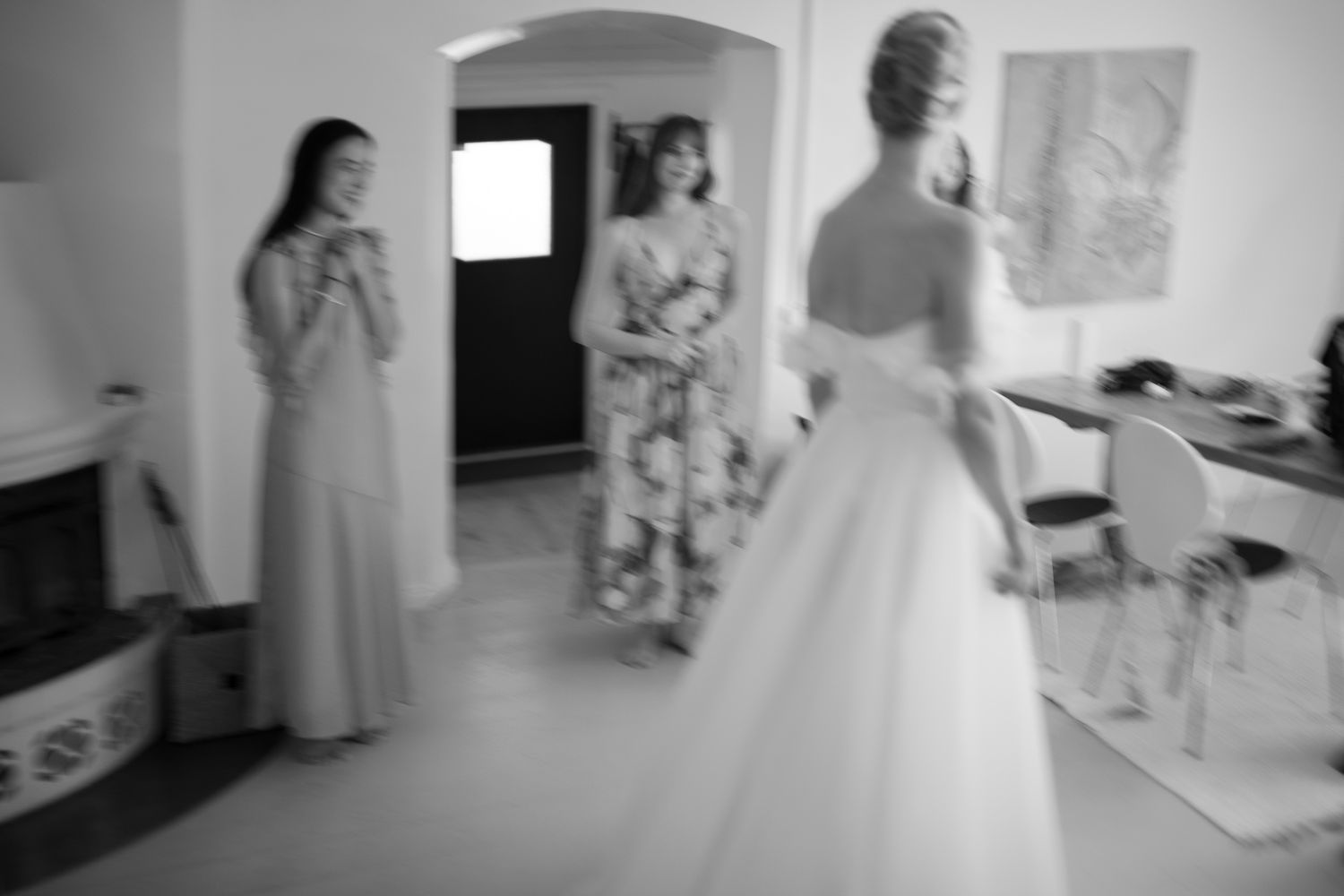 Black and white photo of a bride in a white gown being admired by two bridesmaids in a elegant room.