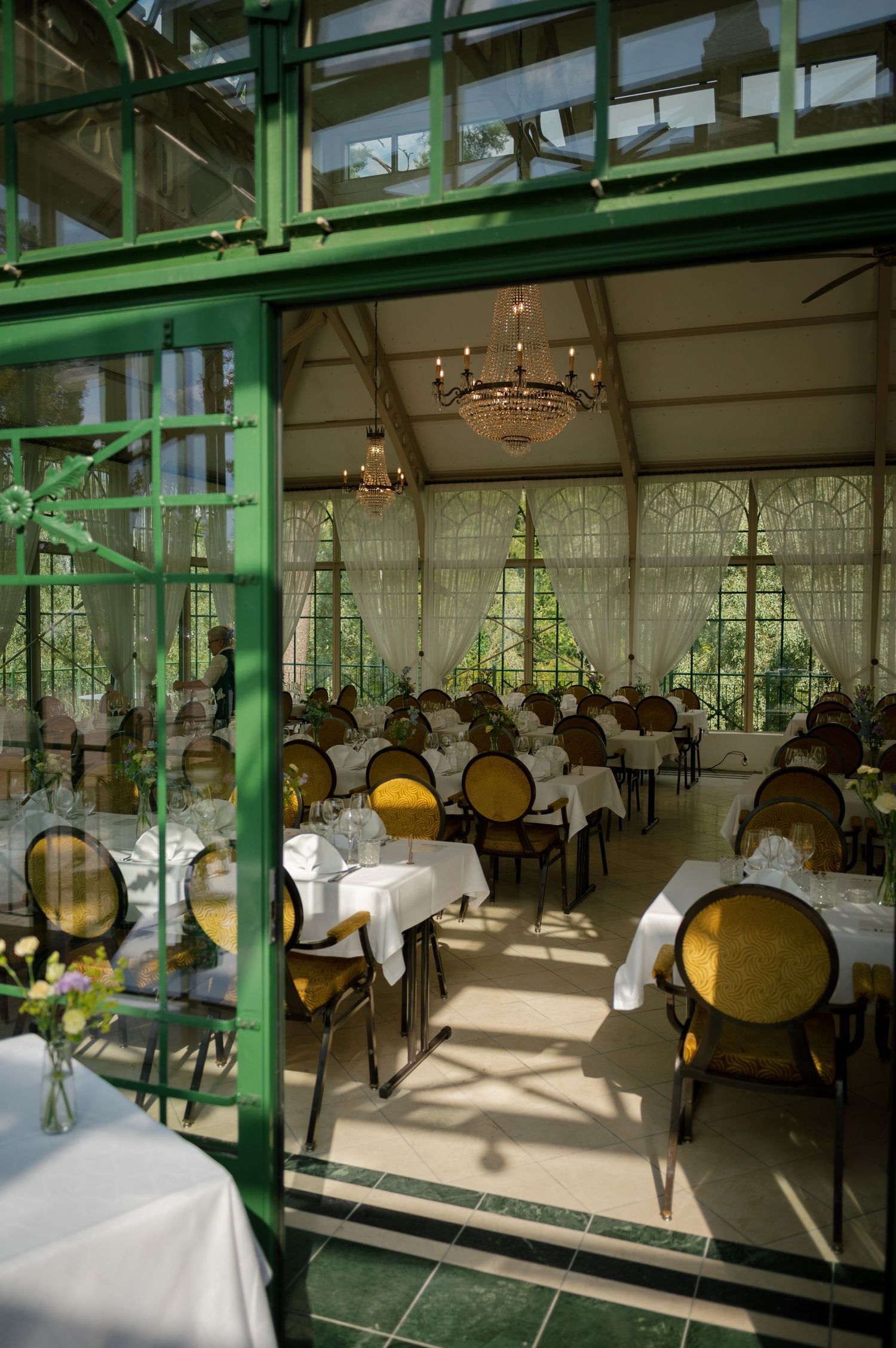 Sunlit restaurant interior with green structural elements, white tablecloths, and classic dining chairs.