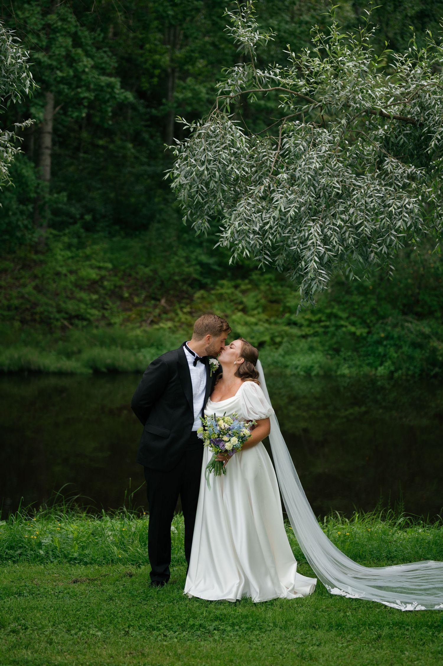 Newlyweds share an intimate moment by a pond with flowing white dress and tuxedo.