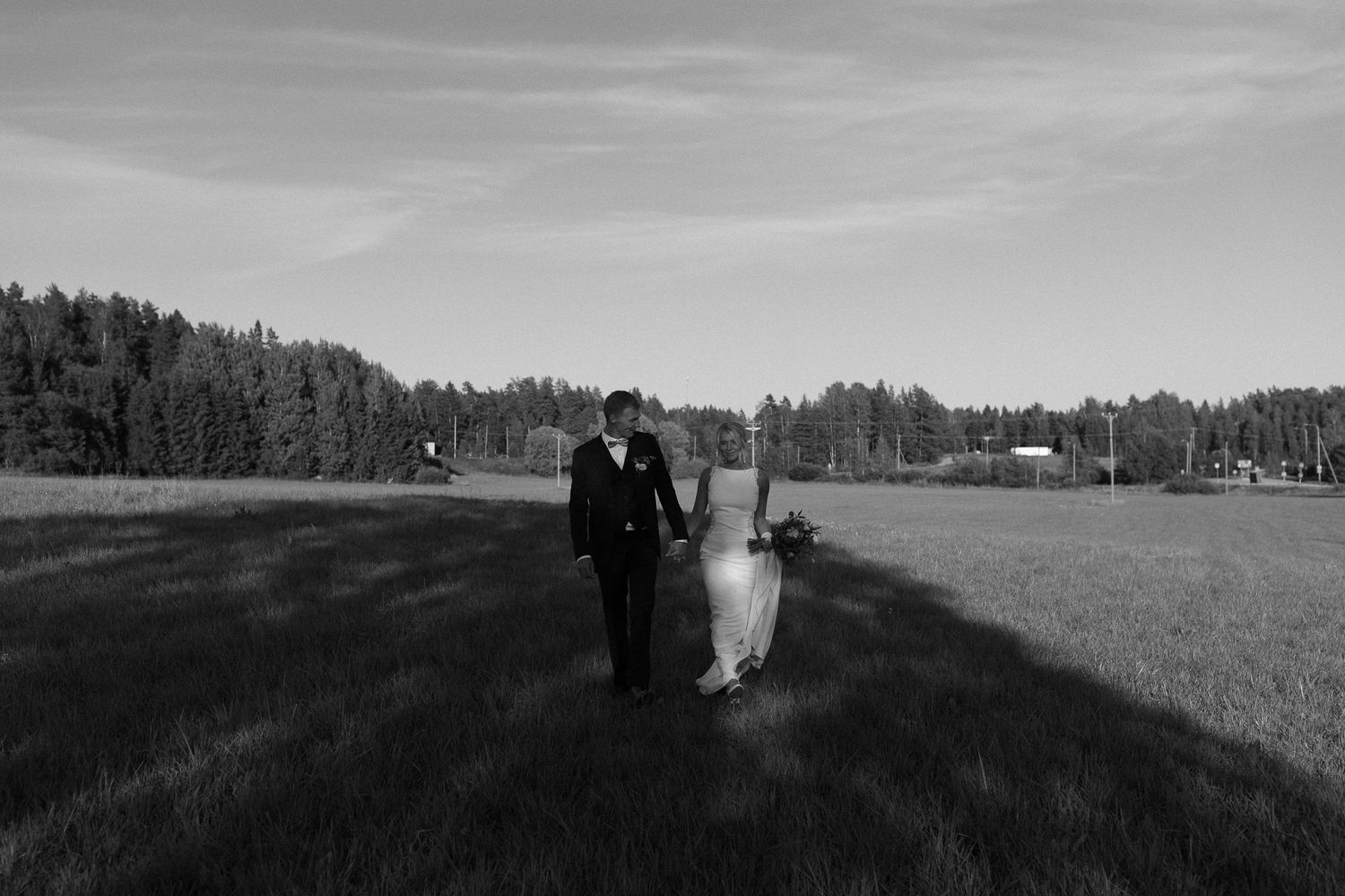 Black and white landscape photo of couple walking through expansive field with forest backdrop.