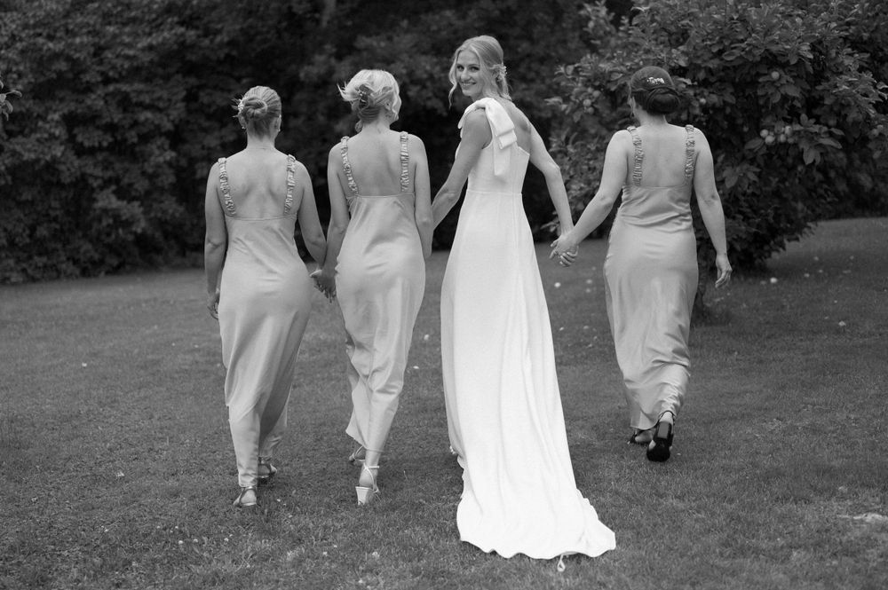 Black and white photo of wedding party walking away across lawn in flowing dresses.