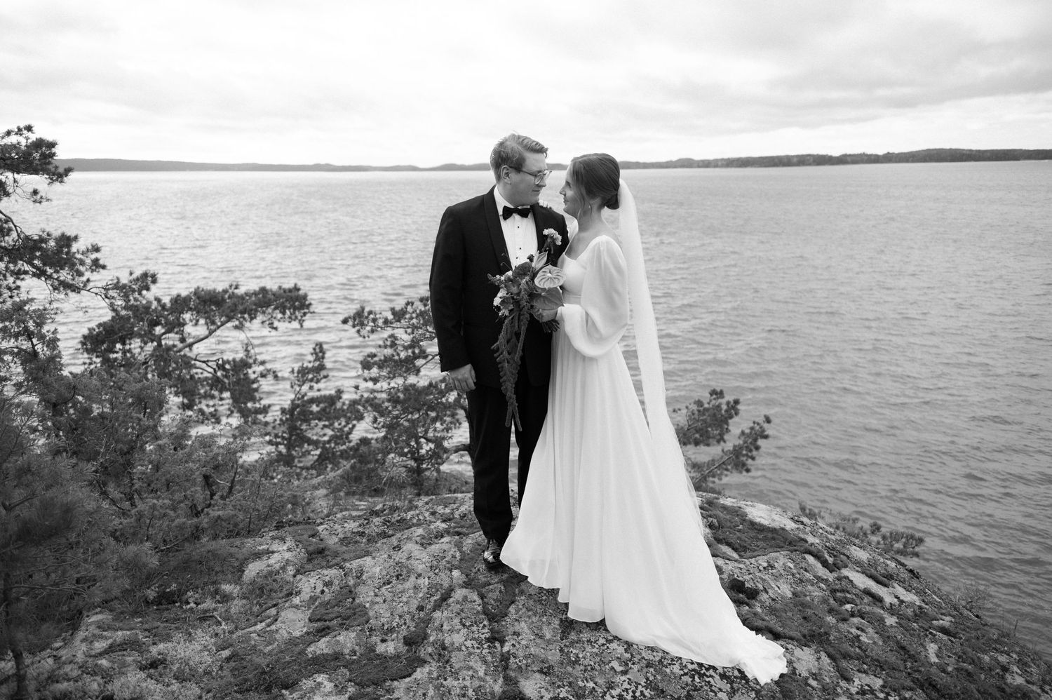 A couple in formal attire stands together on a scenic cliffside overlooking a serene lake at sunset.