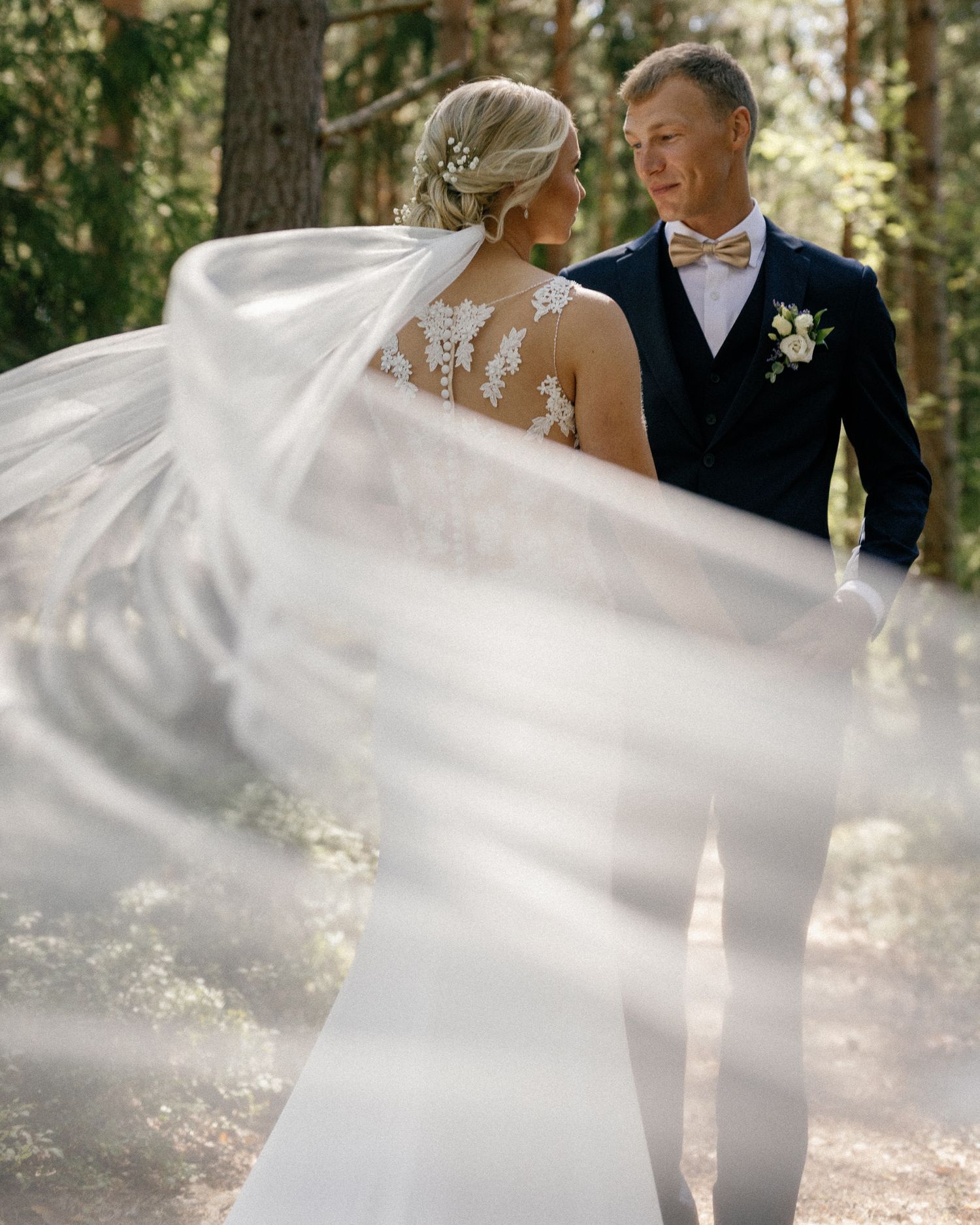 A wedding couple poses in a sunlit forest while the bride's veil flows dramatically in the breeze.